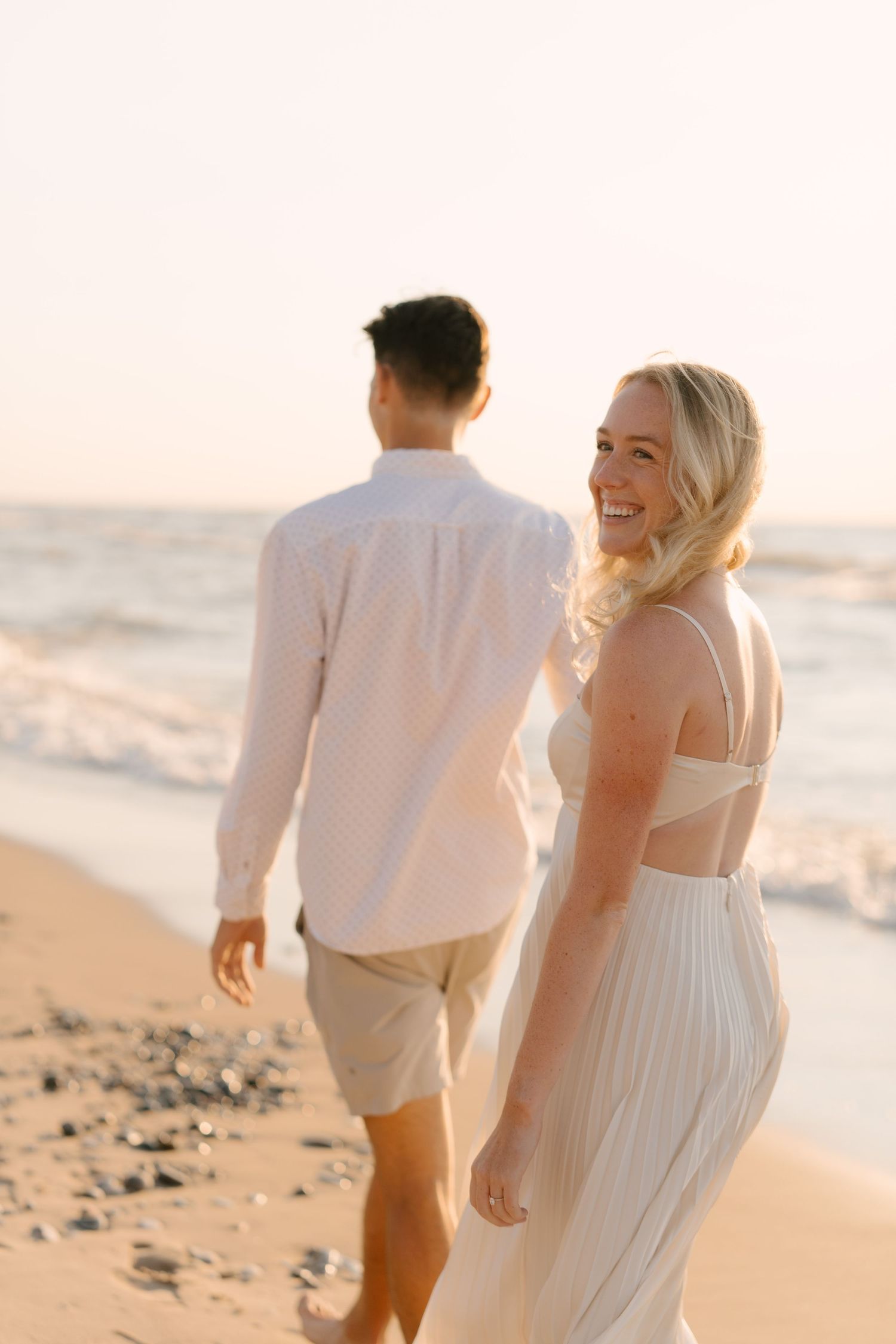 Smiling blonde woman in white dress walks with partner along sunlit beach at sunset.