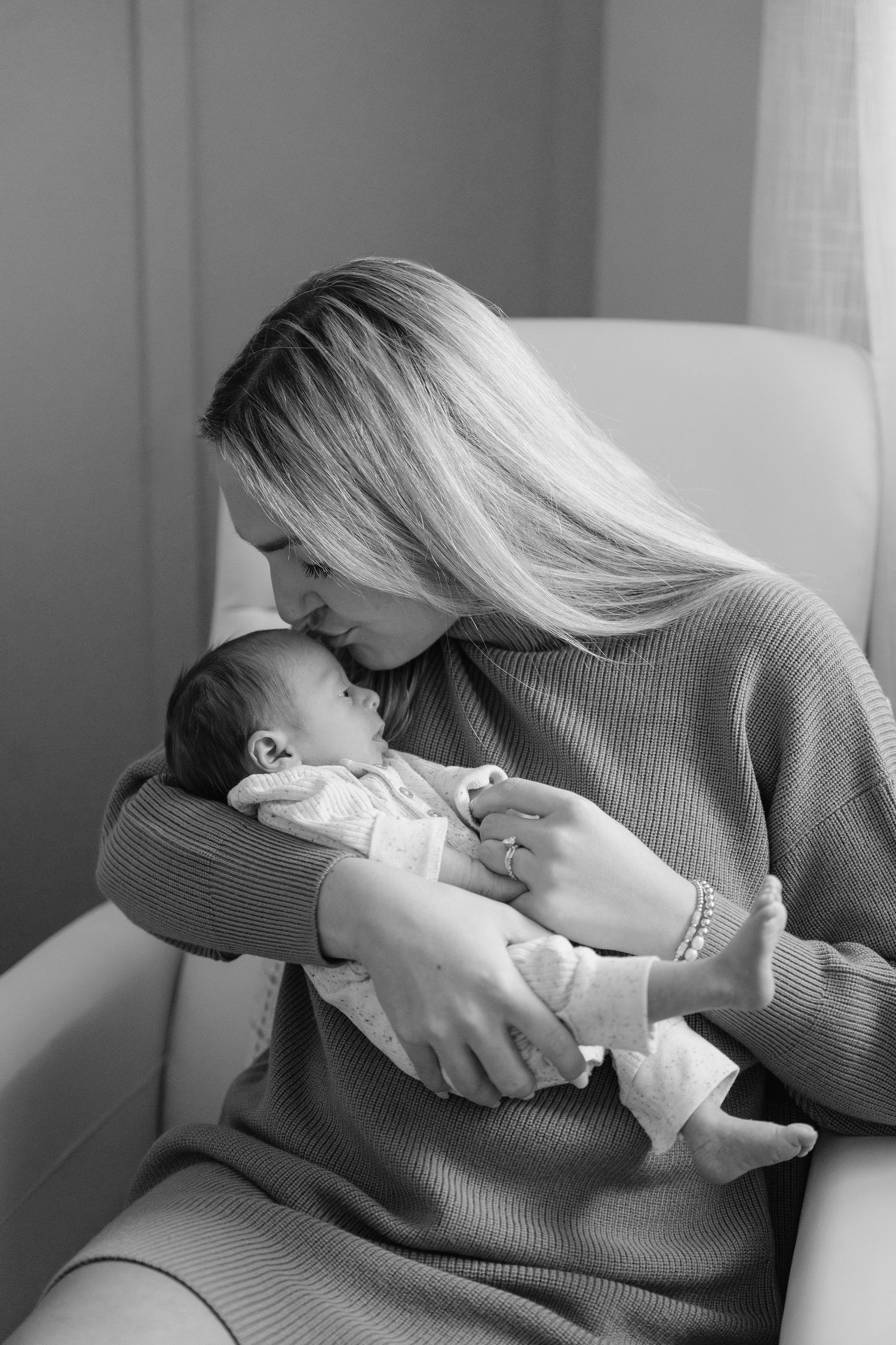 Black and white photo of loving mother kissing her newborn baby while seated in chair.