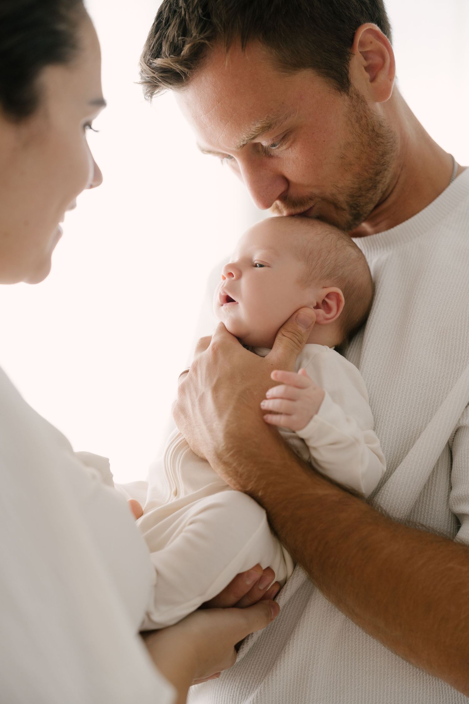 Father kissing newborn baby on forehead while mother looks on in bright natural light.