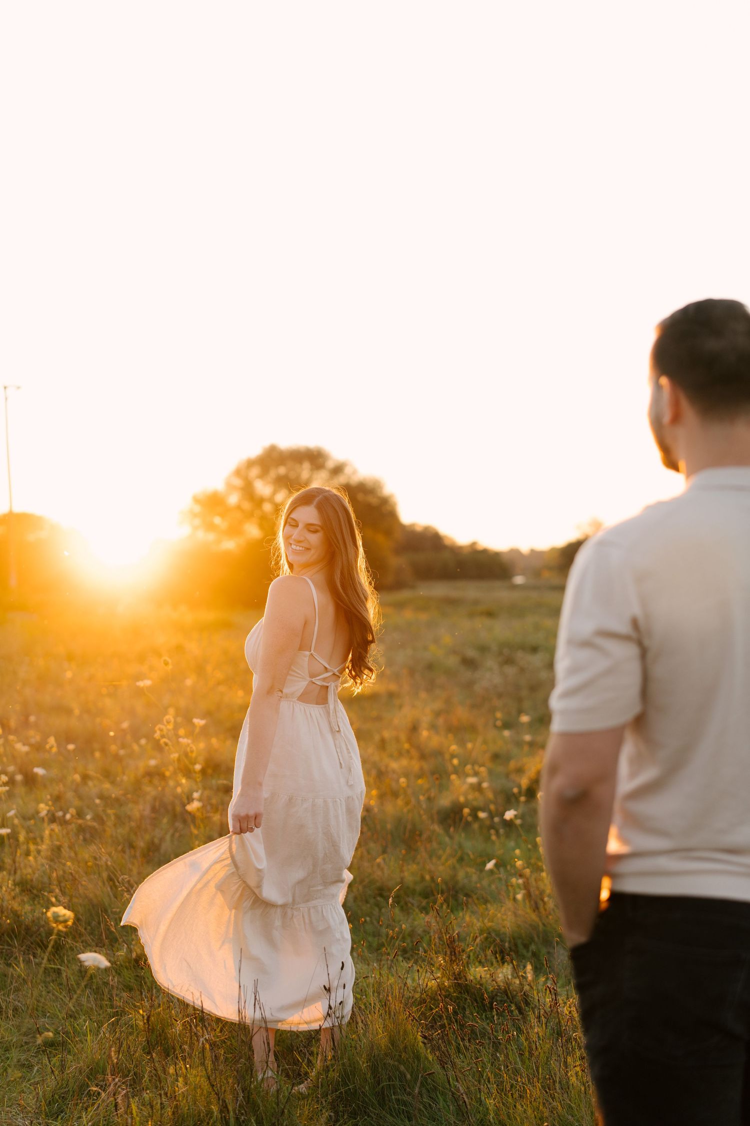 Woman in white dress smiling back at her partner in a glowing golden sunset field.