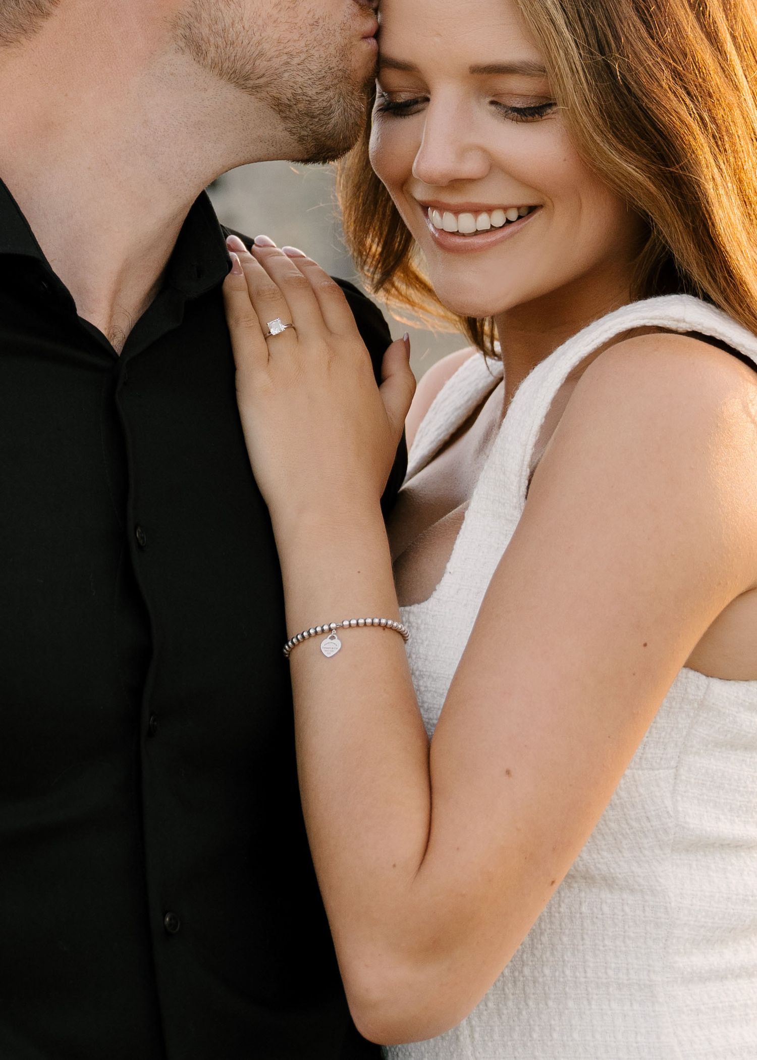 Close-up of woman smiling with engagement ring as man kisses her temple.