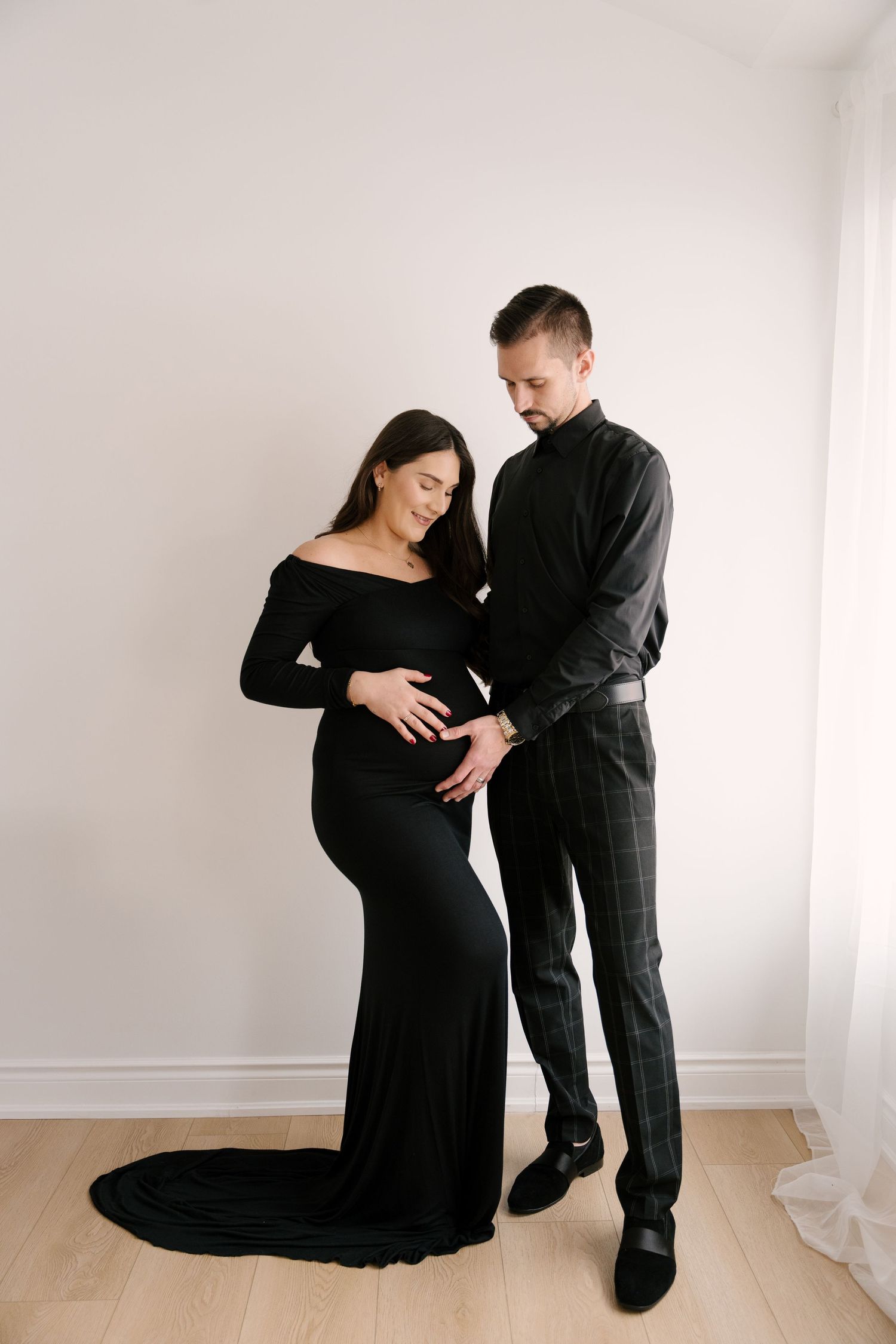 Elegant pregnant woman in black off-shoulder gown with partner in black outfit admiring her baby bump.