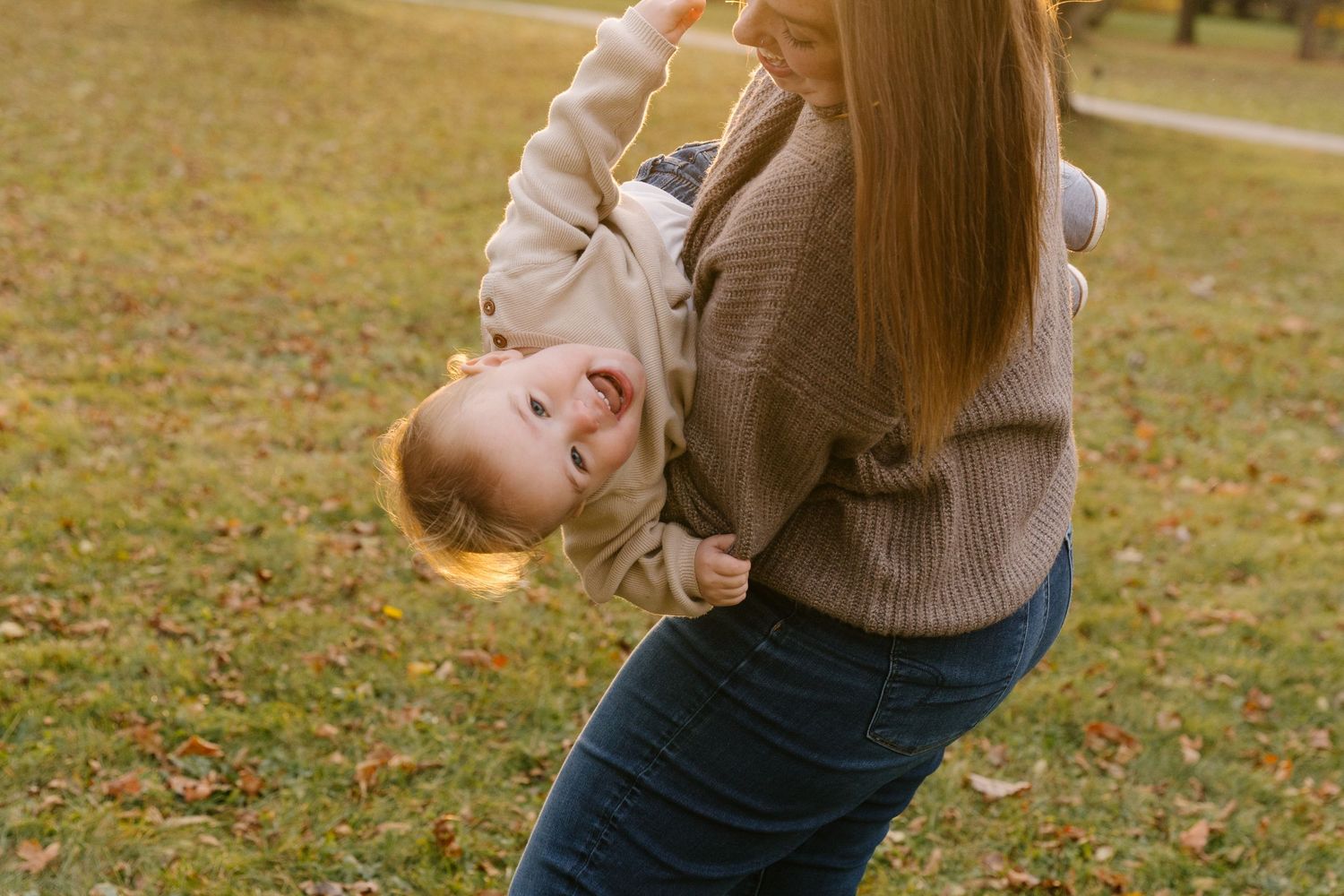Playful mother in brown sweater swinging laughing toddler upside down in fall park.
