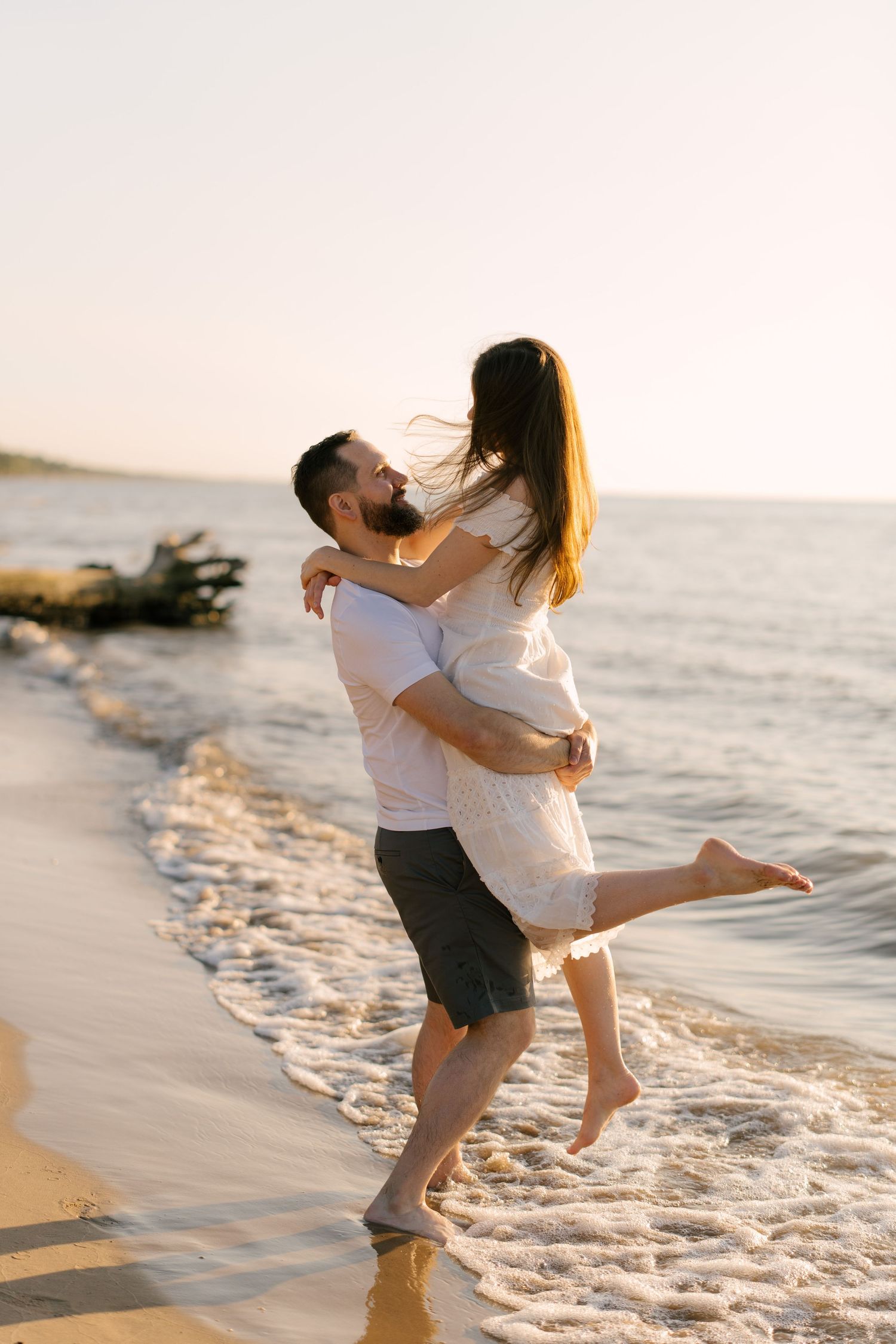 Romantic couple on a sandy beach at sunset, man lifting woman in white dress while ocean waves wash ashore.