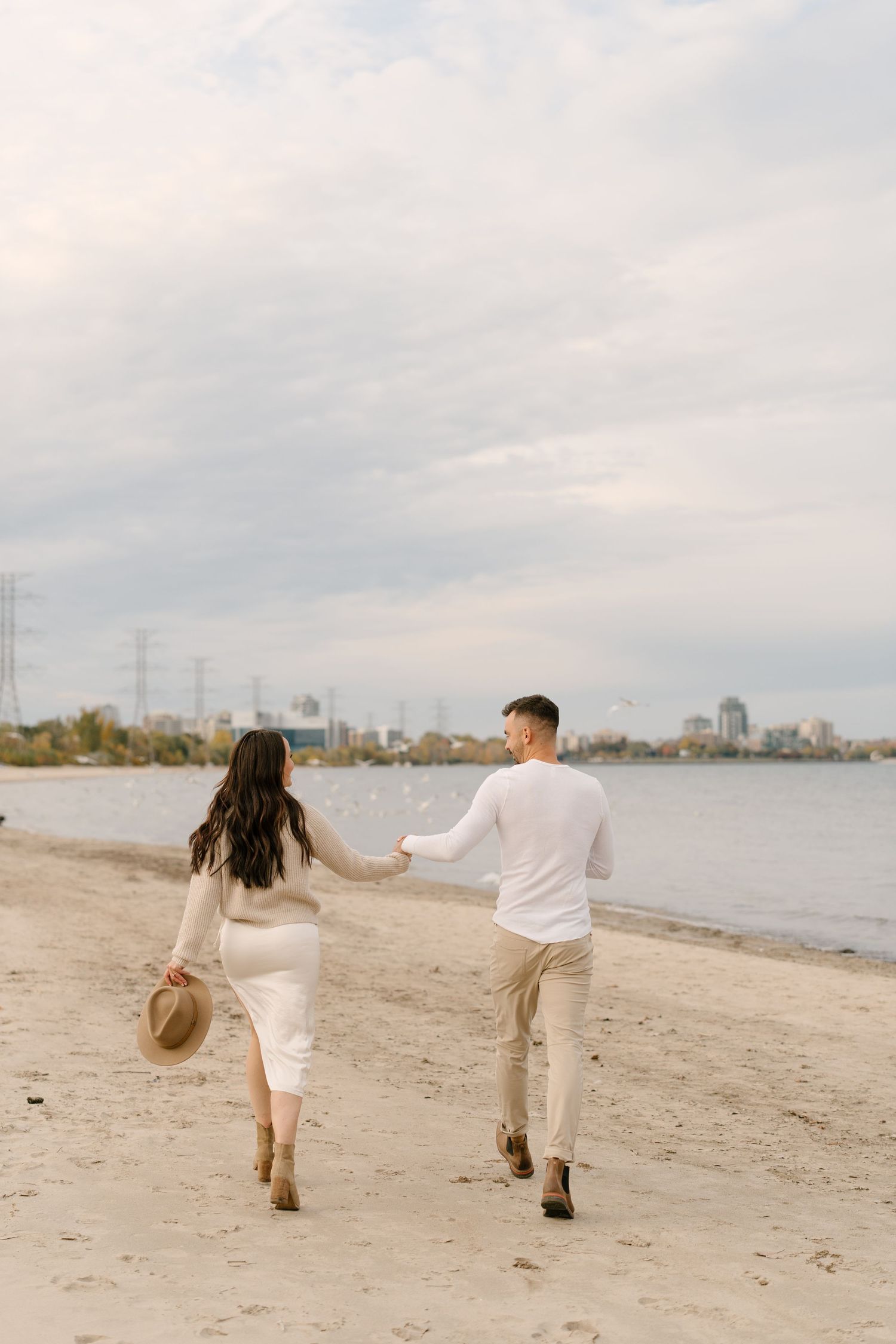 Couple holding hands walking along sandy beach shoreline under cloudy sky.