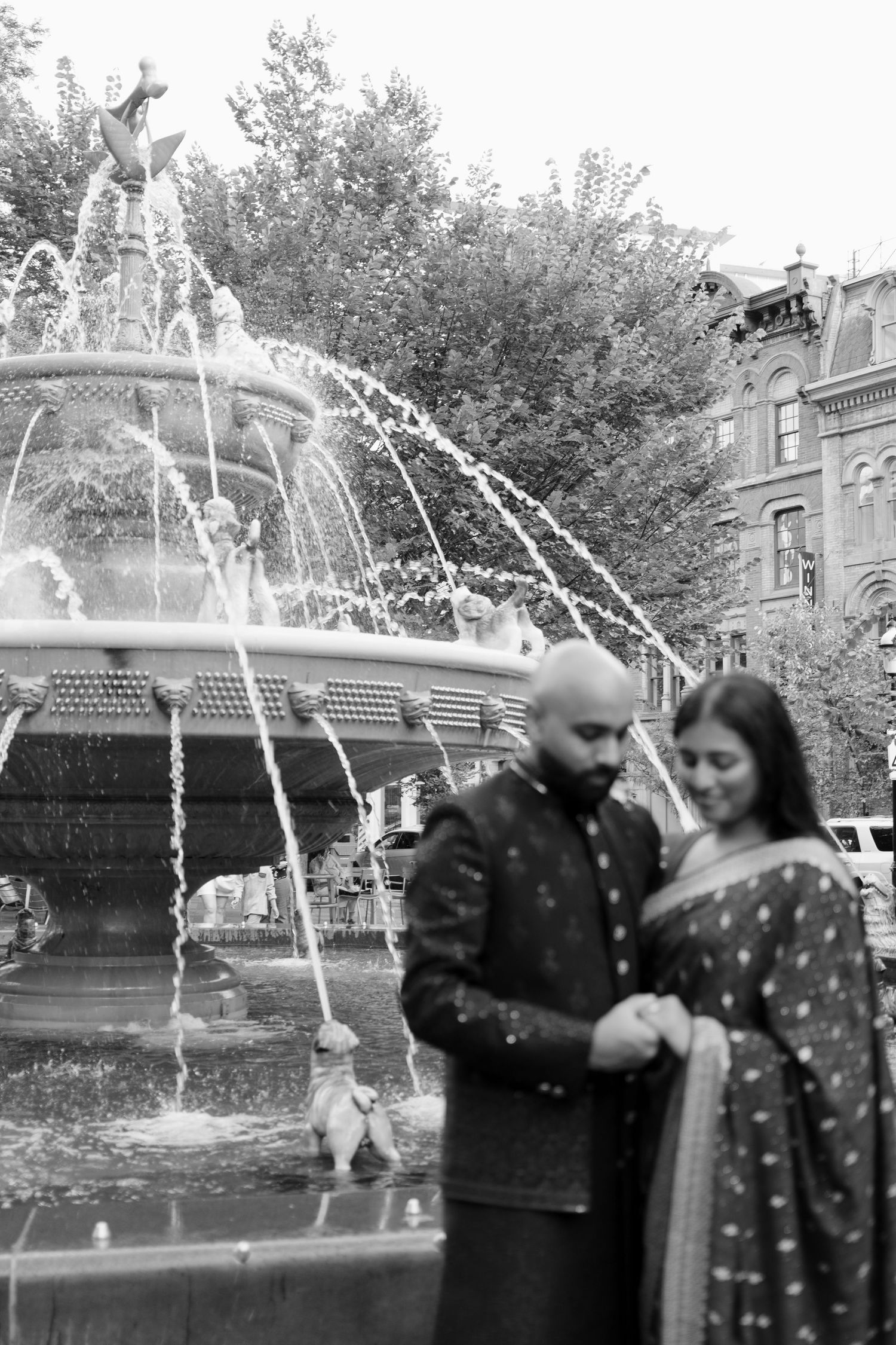 Indian couple in traditional attire share a tender moment near an ornate city fountain in Toronto.