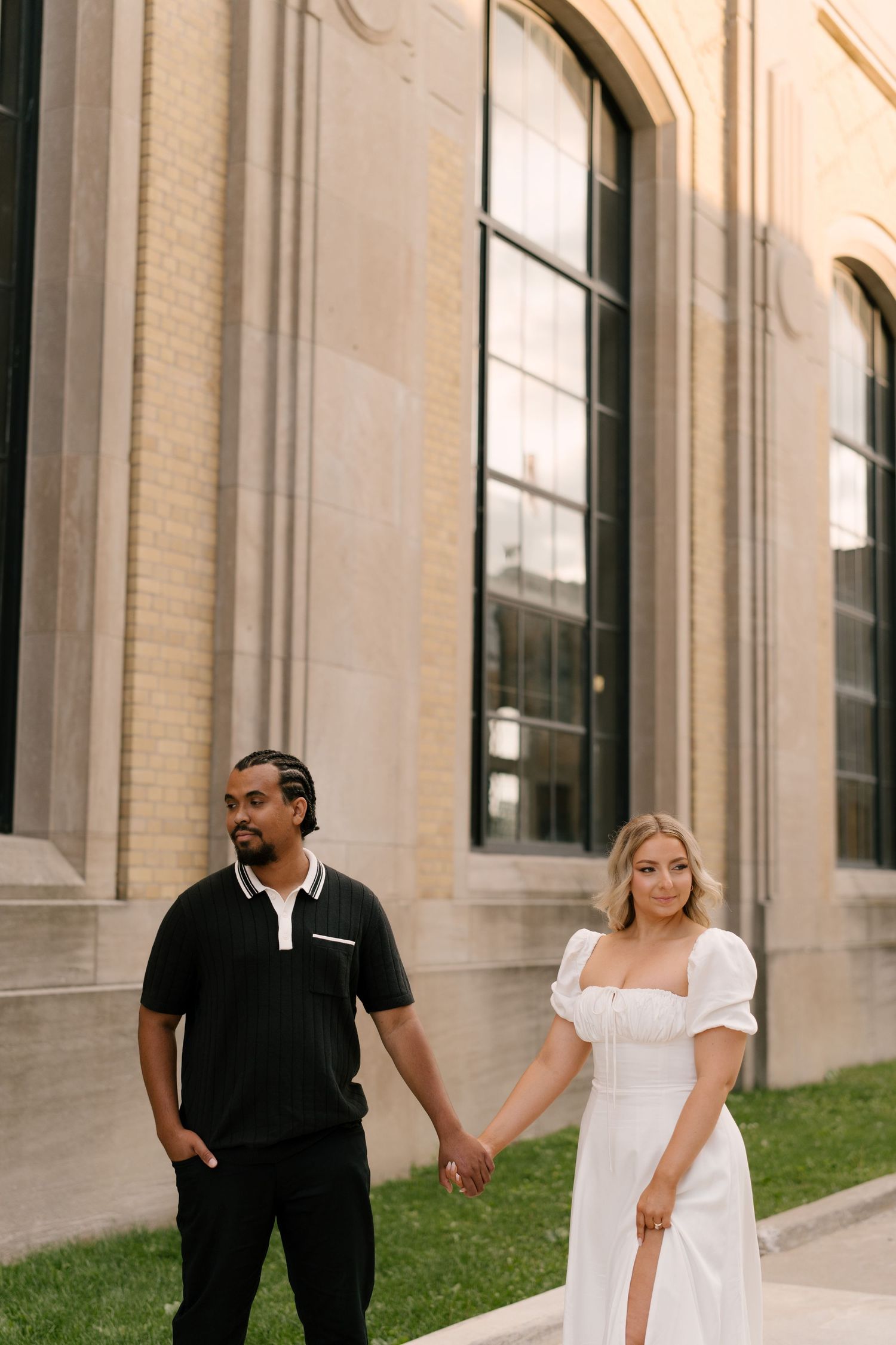 Couple holding hands walking past large arched windows of a classic stone building on sunny day.