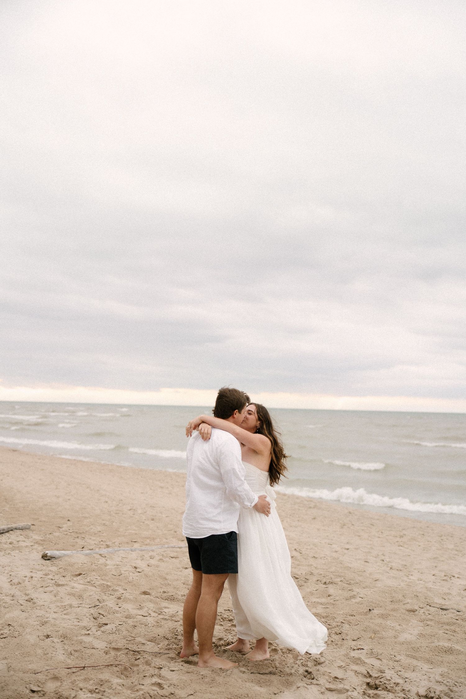 Couple sharing a romantic embrace on a sandy beach with ocean waves and cloudy sky behind them.