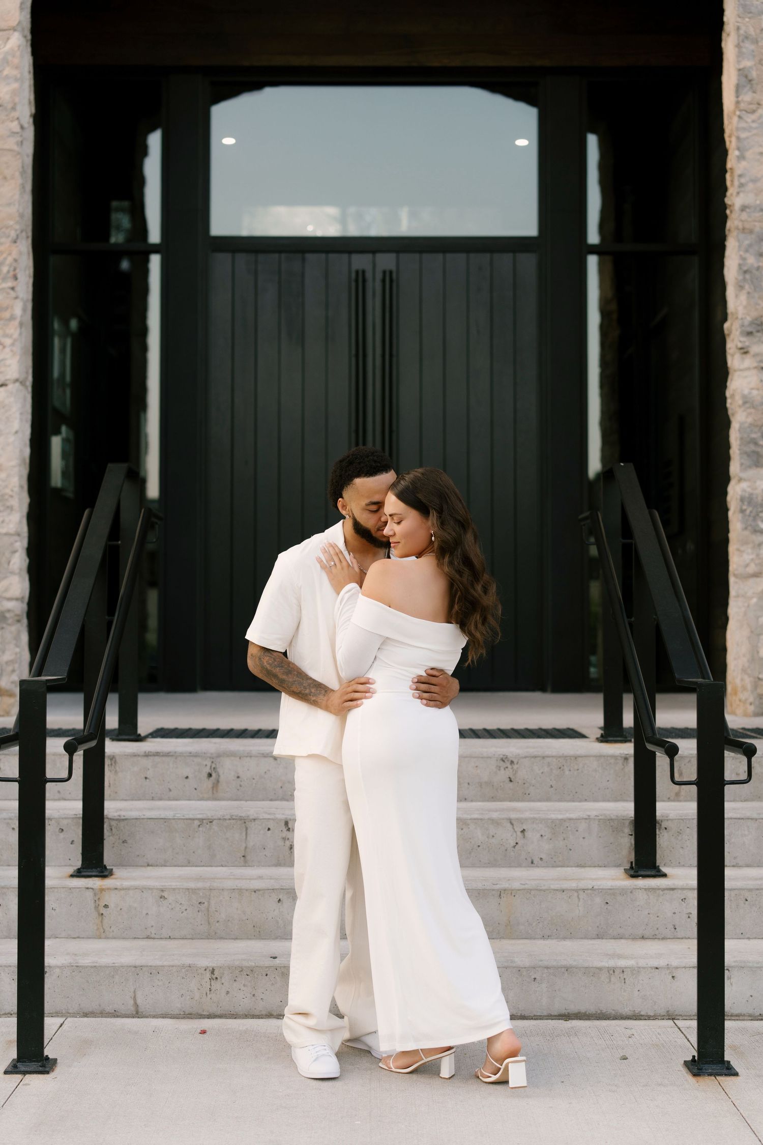 Couple in elegant white outfits embrace before black double doors on stone staircase.