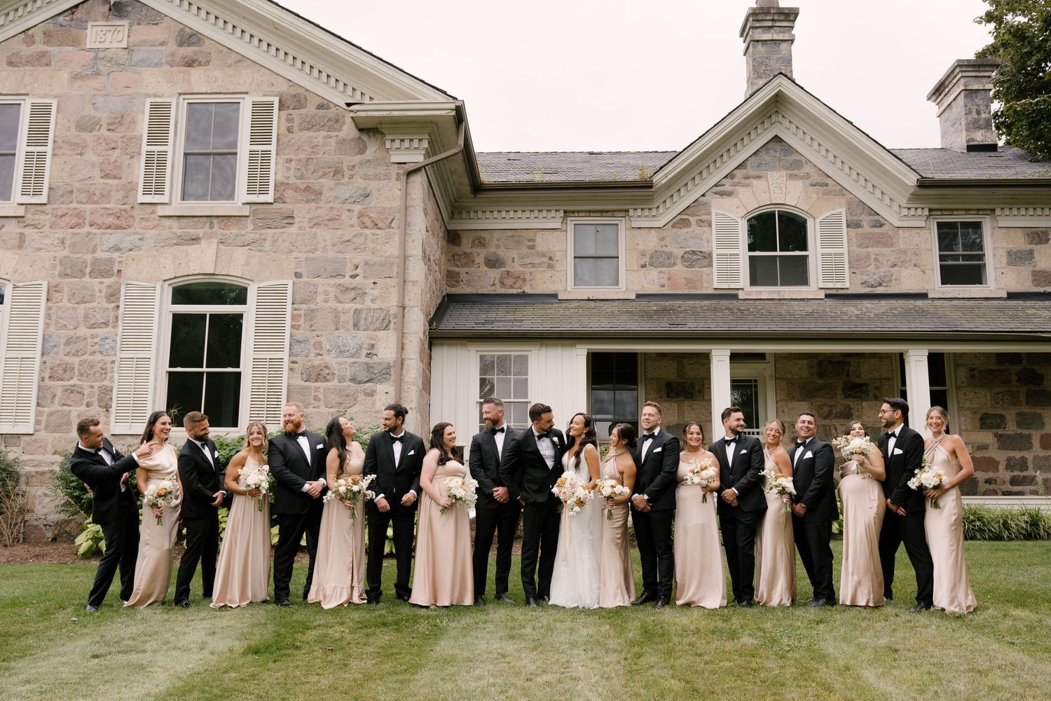 Large wedding party standing together on lawn in front of a beautiful historic stone Wakefield Estate.