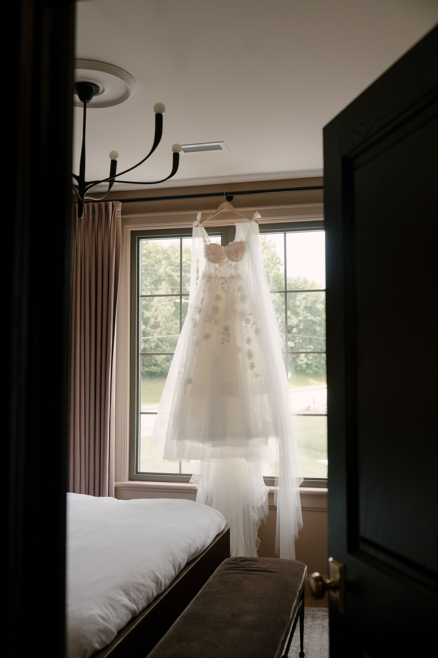 Floral applique wedding gown hanging in a sunlit bedroom window before the ceremony.