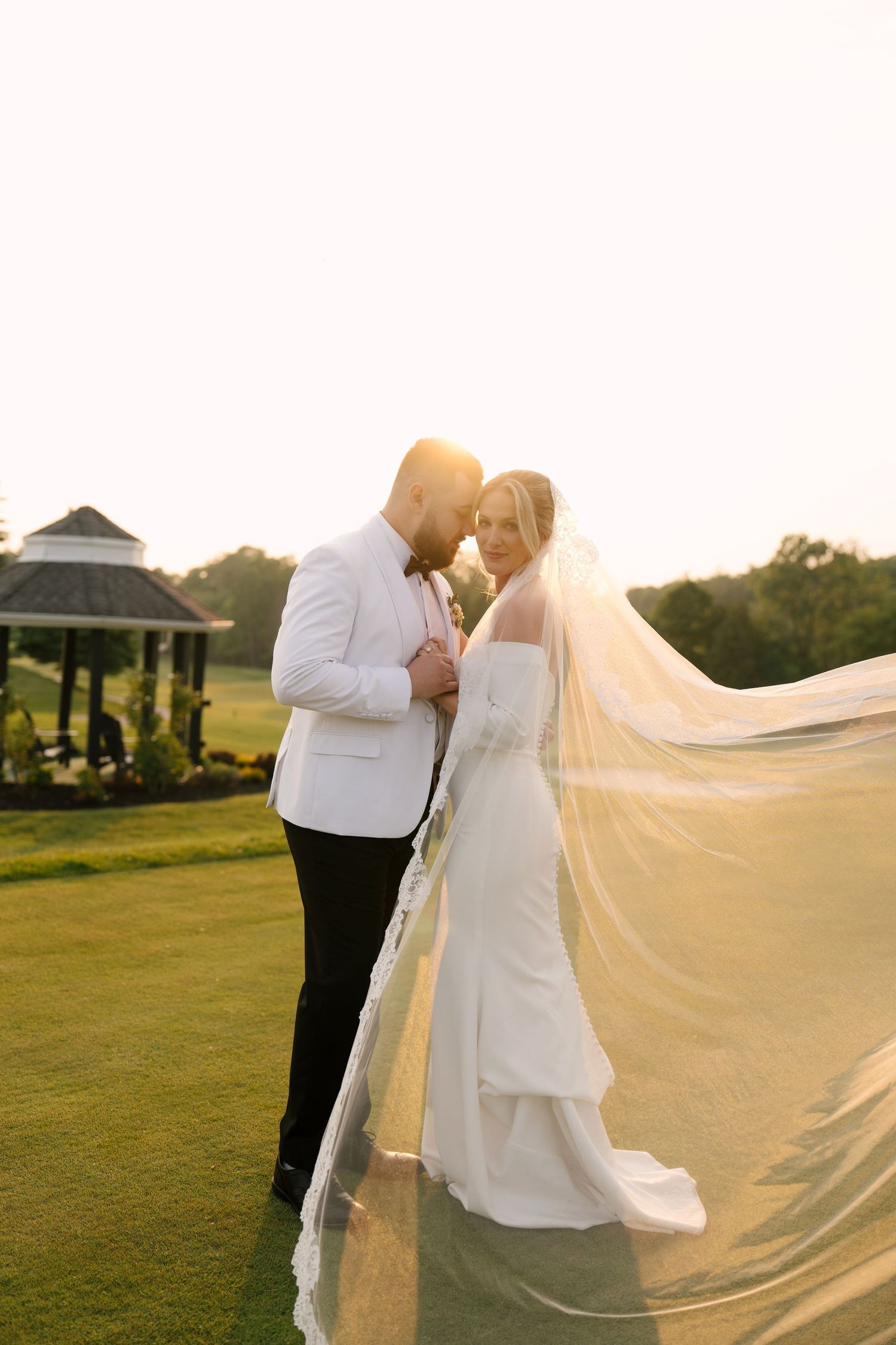 Bride and groom standing close at sunset on Whistle Bear golf course, cathedral lace veil flowing dramatically in the wind.