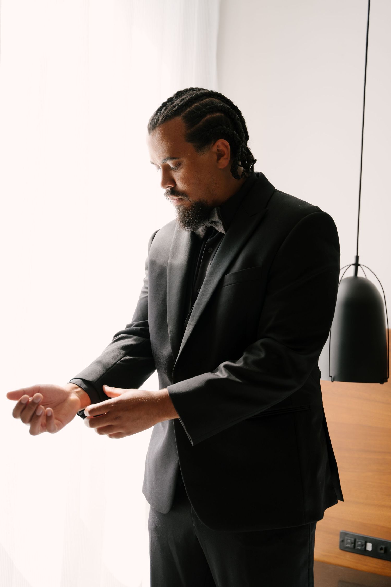Man in black suit adjusting his watch cufflinks near a bright window.