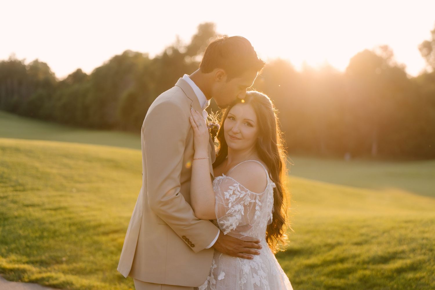 Groom in tan suit tenderly embraces brunette bride in lace gown during golden sunset at Whistle Bear wedding venue.