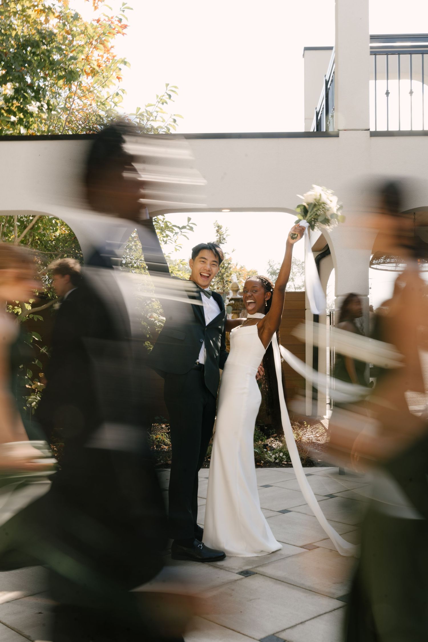 Joyful interracial newlyweds celebrate as guests pass by in a lively motion-blurred wedding photo at Wren House.