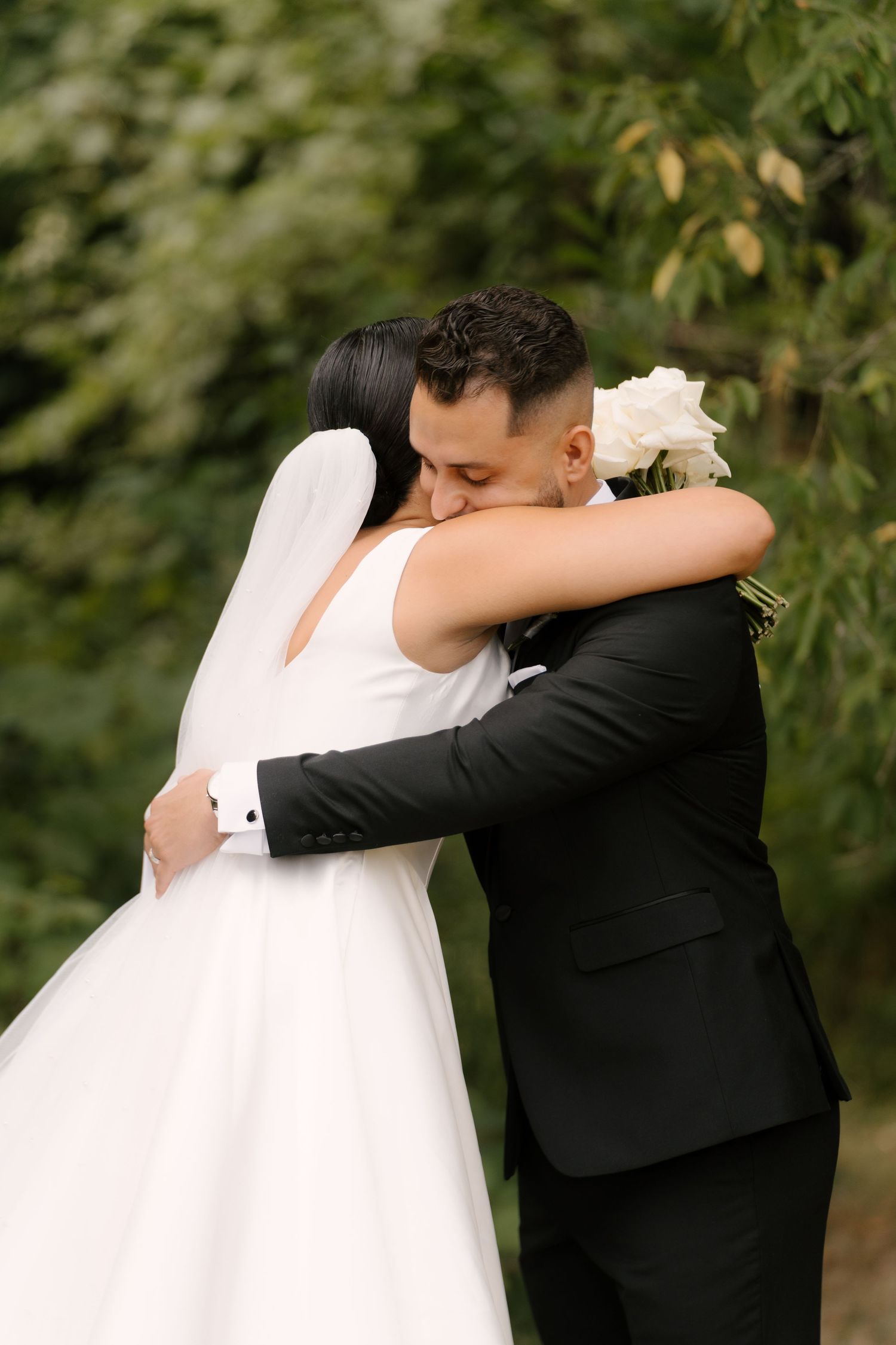 Bride and groom share tender embrace outdoors, bride holding white bouquet against lush green garden backdrop.
