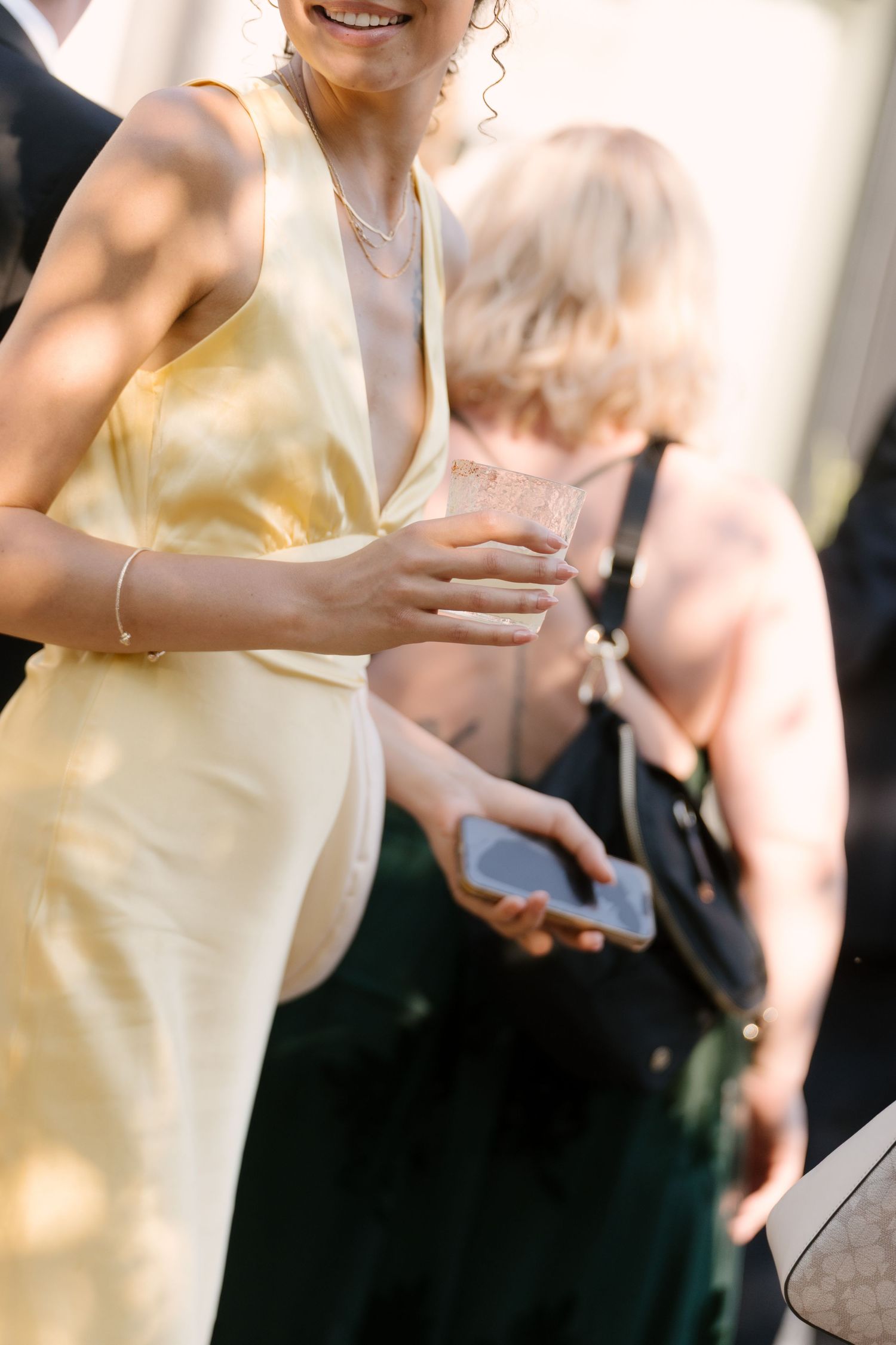 Wedding guest in yellow satin halter dress holding champagne glass and smartphone at reception.