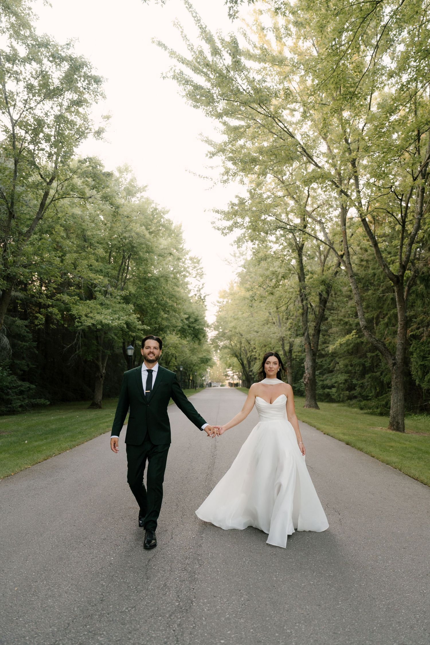 Bride and groom holding hands walking down a tree-lined road, bride in white strapless gown, groom in dark suit at Roseville Estates.
