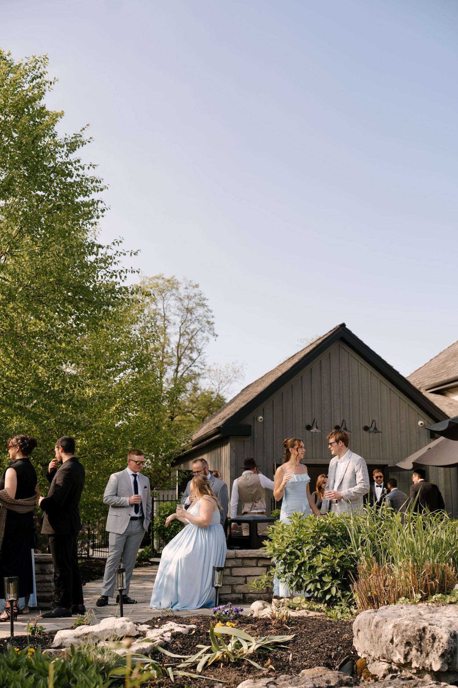 Elegant outdoor wedding reception with guests in formal attire gathered near a rustic venue surrounded by lush greenery at Roseville Estates