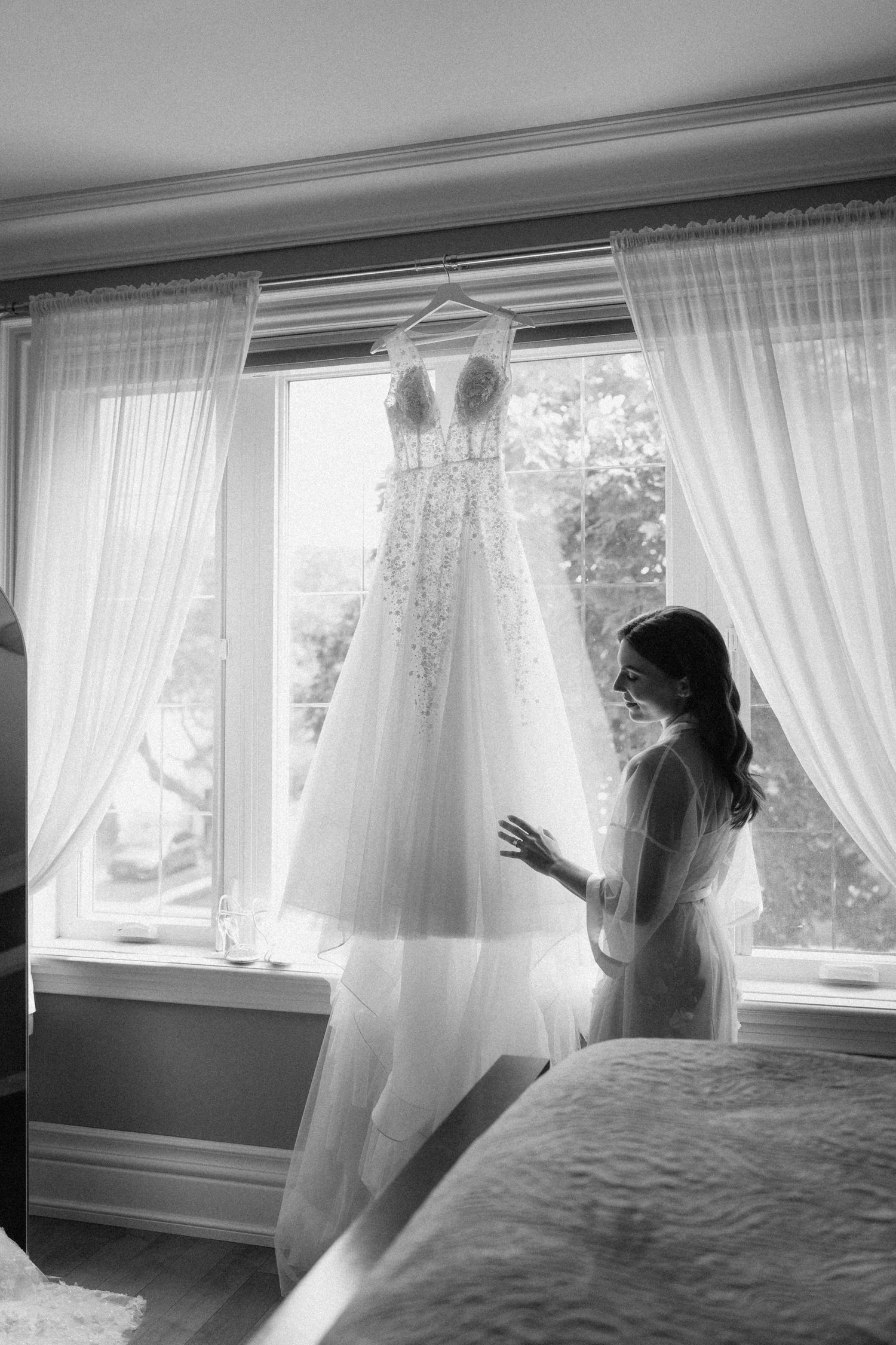 Black and white photo of bride admiring her lace wedding gown hanging in a sunlit window.