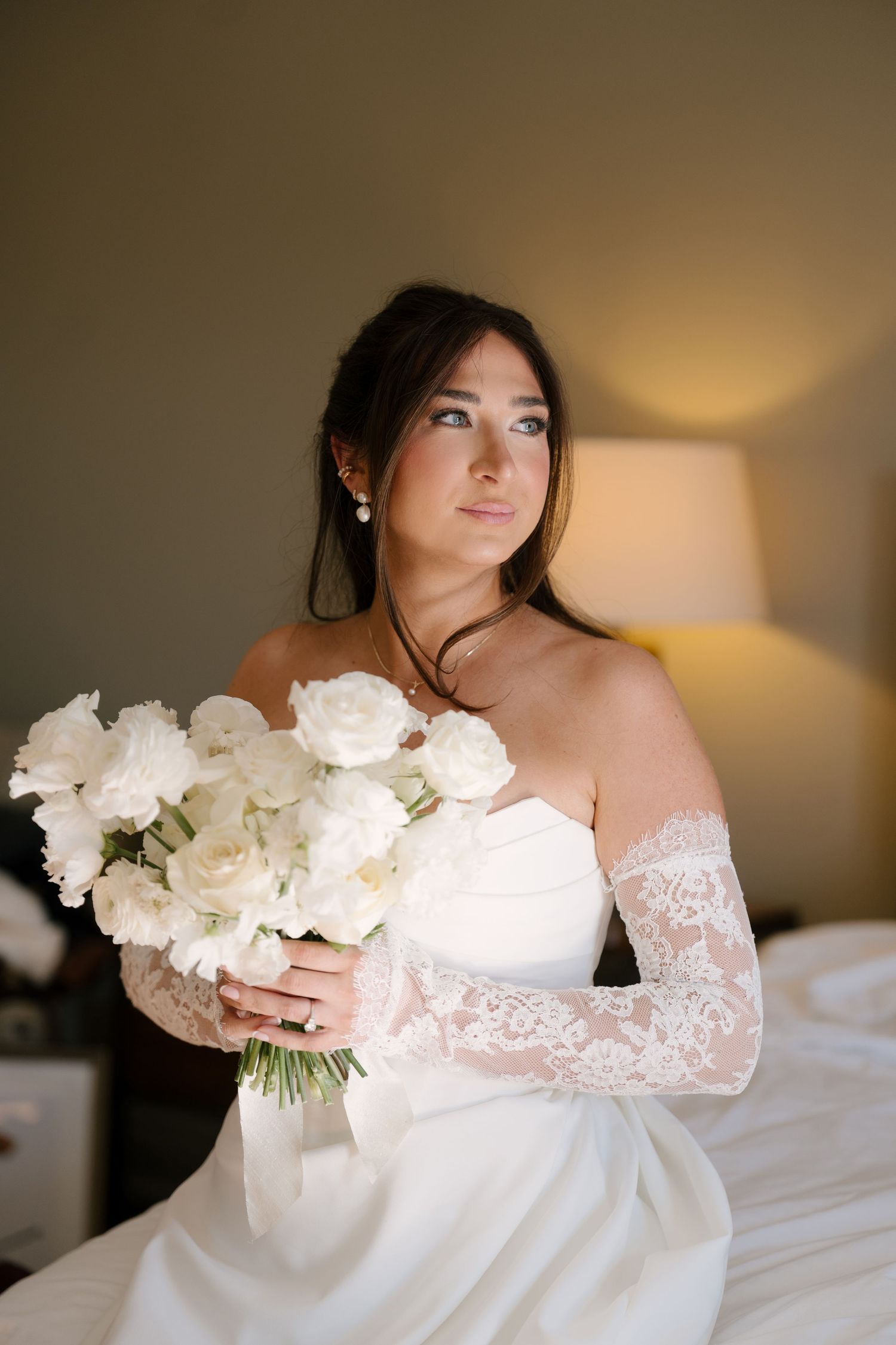 Brunette bride in strapless white gown with lace gloves holds white peony bouquet indoors.