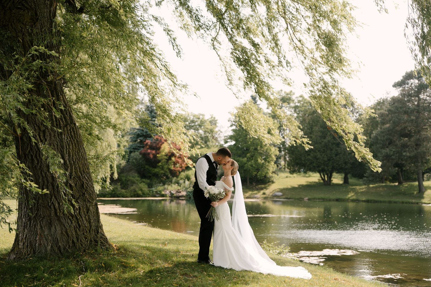 Wedding couple embraces near a tranquil lake under a large willow tree in a garden at Guelph golf club.