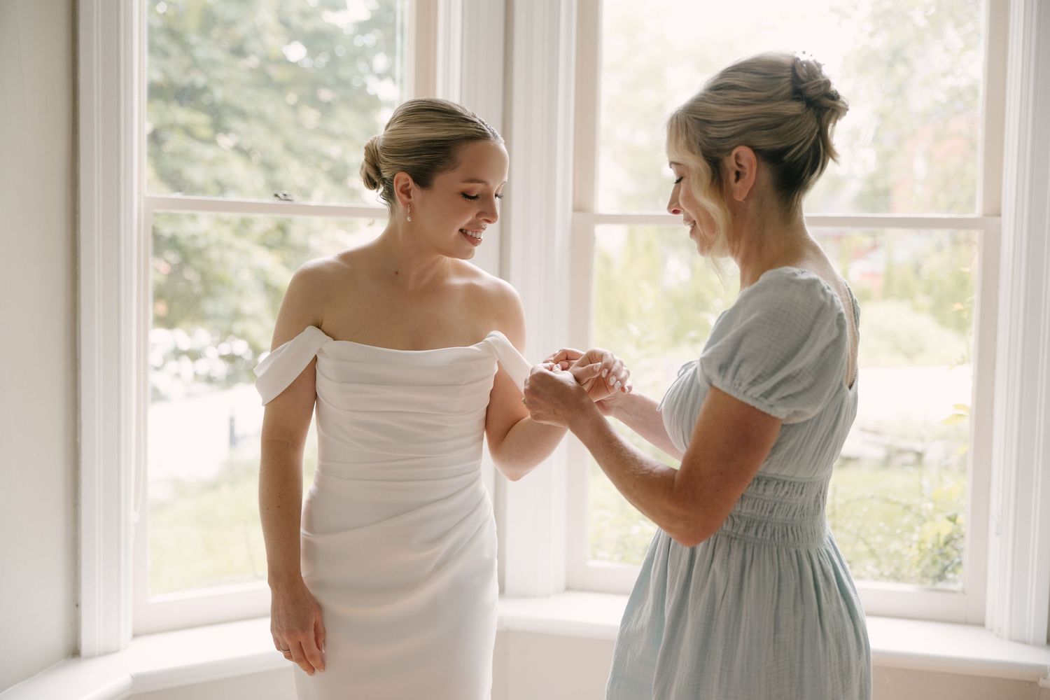 Bride smiling as bridesmaid helps fasten bracelet in bright window light.