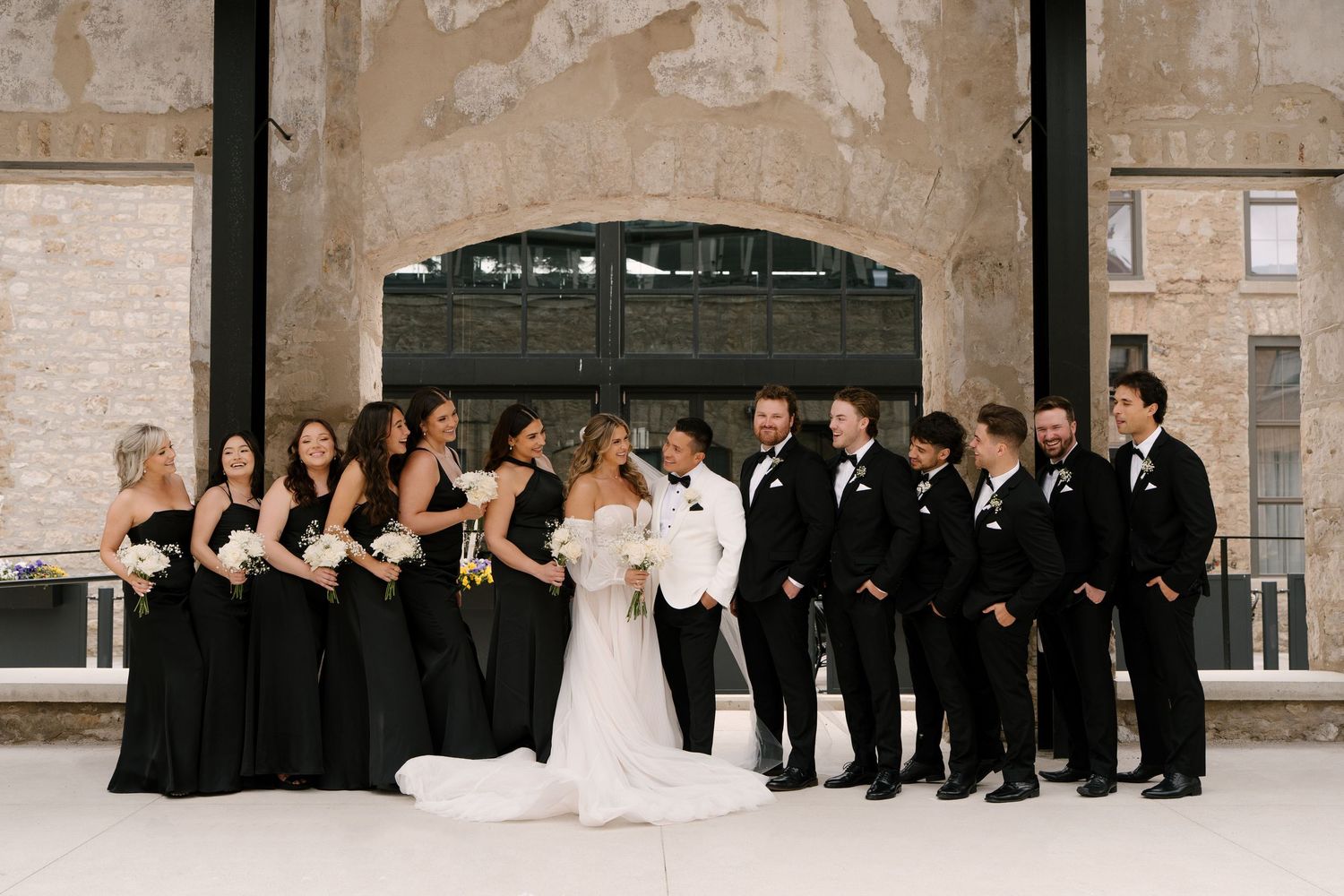 Wedding party in black and white attire poses together outside stone and glass at Tapestry Hall wedding venue.