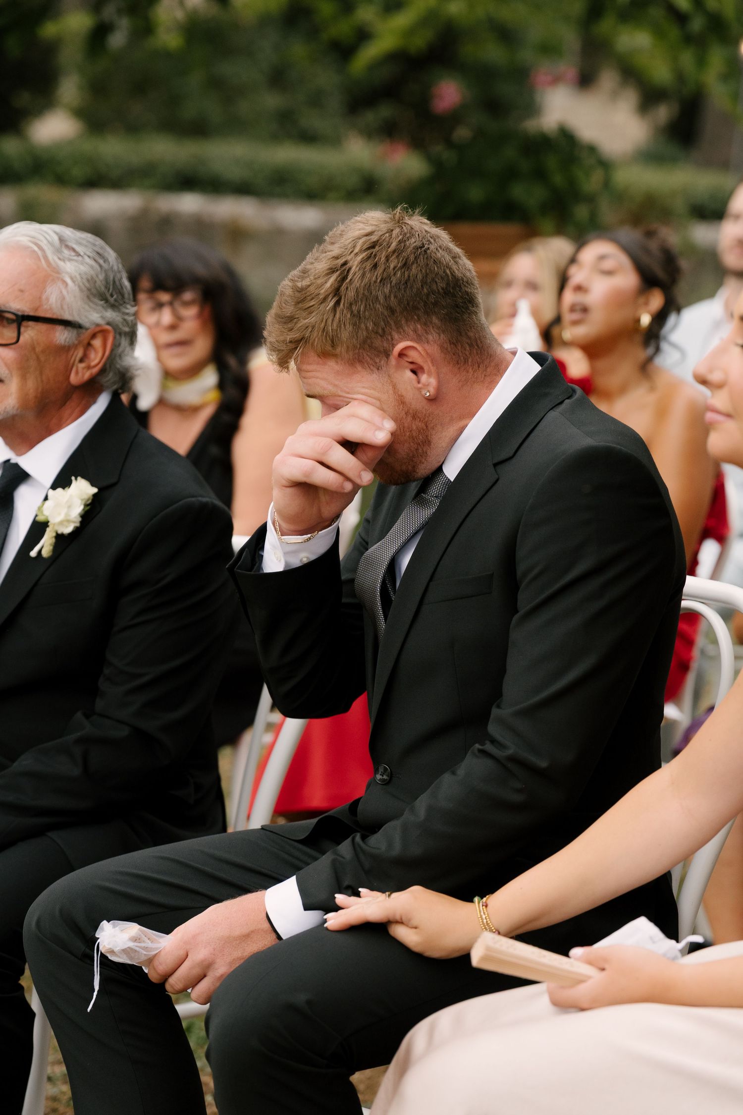 Emotional groom wiping tears at outdoor wedding ceremony, seated in black suit among guests.