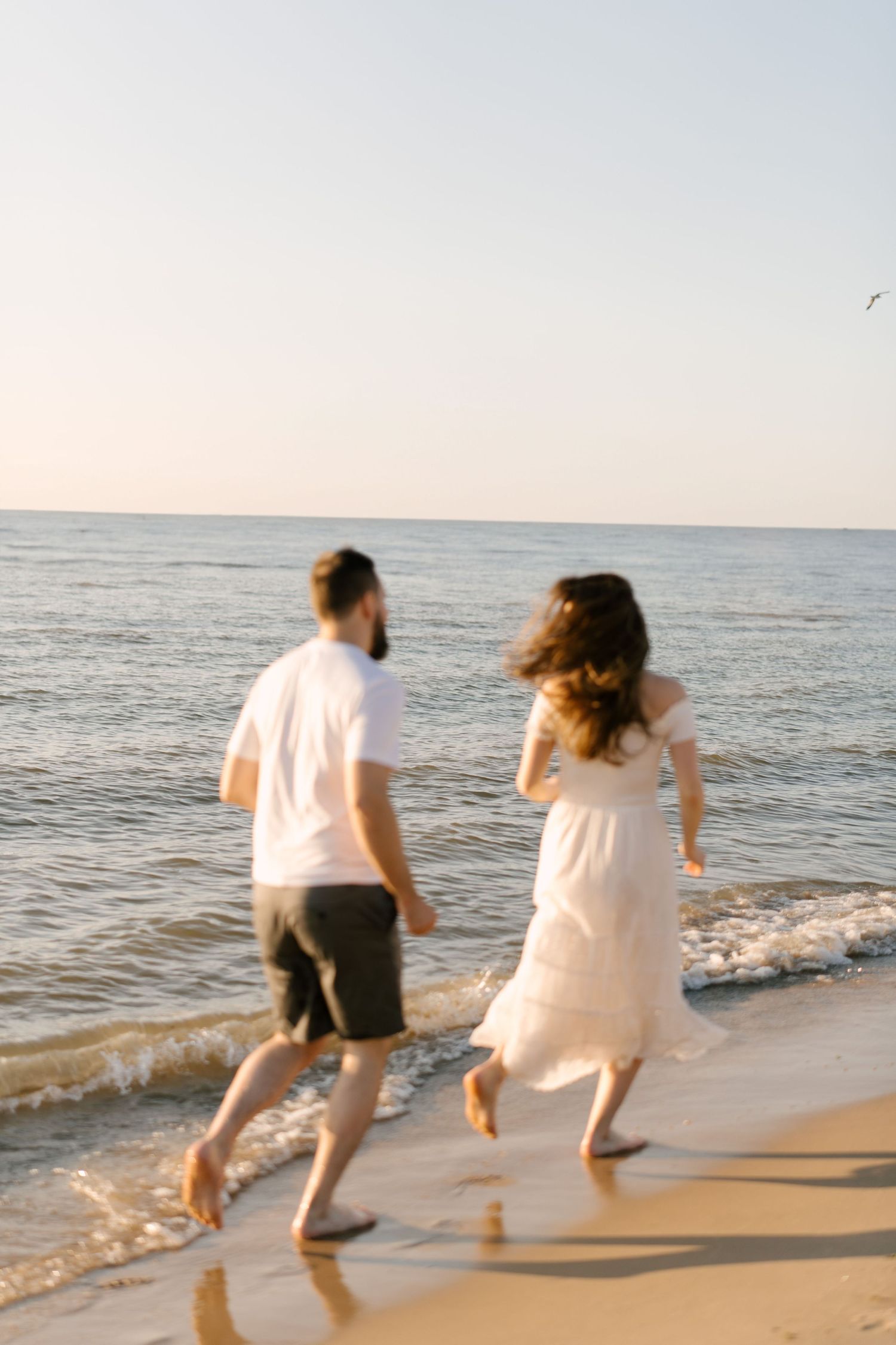 Couple running barefoot along the shoreline at sunset, woman in white dress and man in casual beach attire.