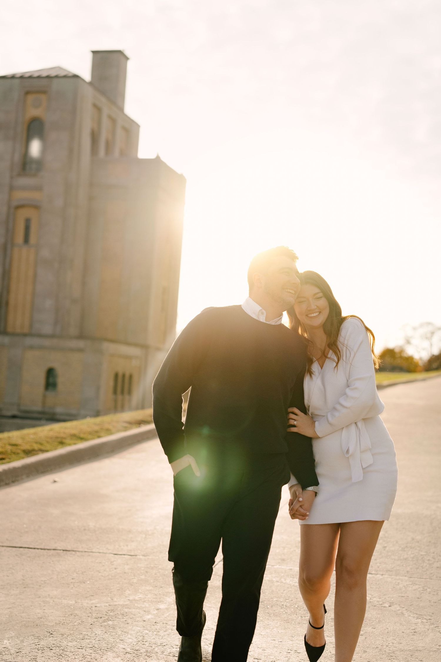 Couple walking together laughing with bright golden sunset and historic building backdrop.