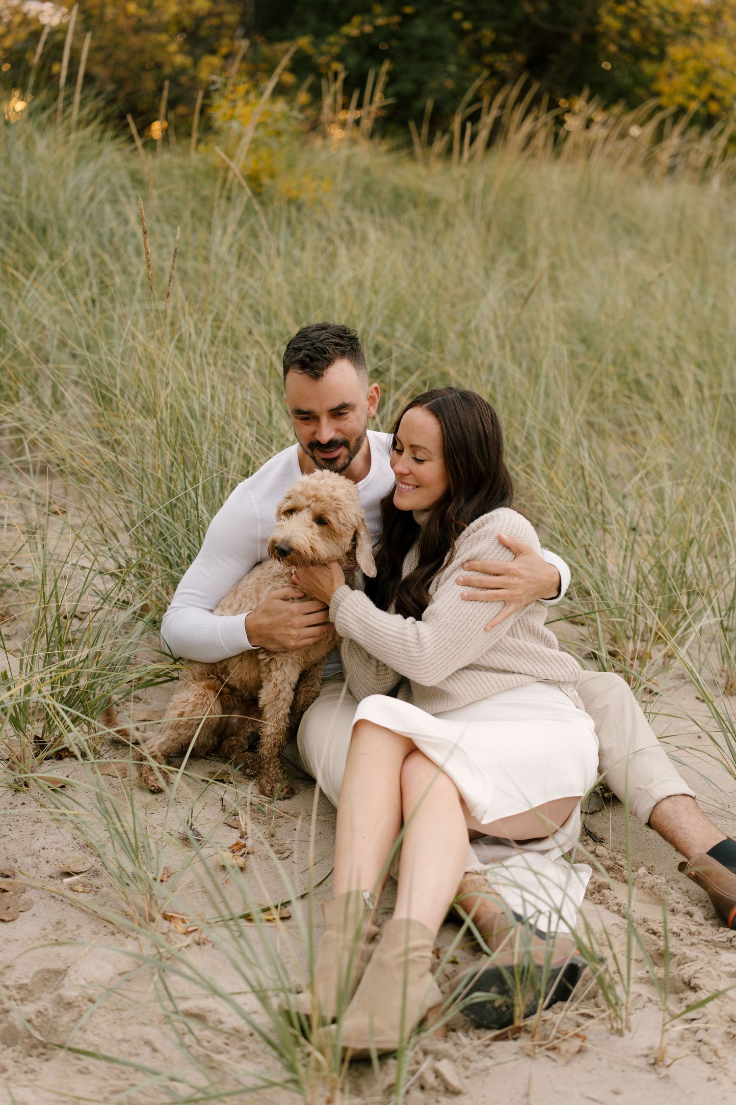 Couple sitting in sand dunes with fluffy goldendoodle dog among tall grass.