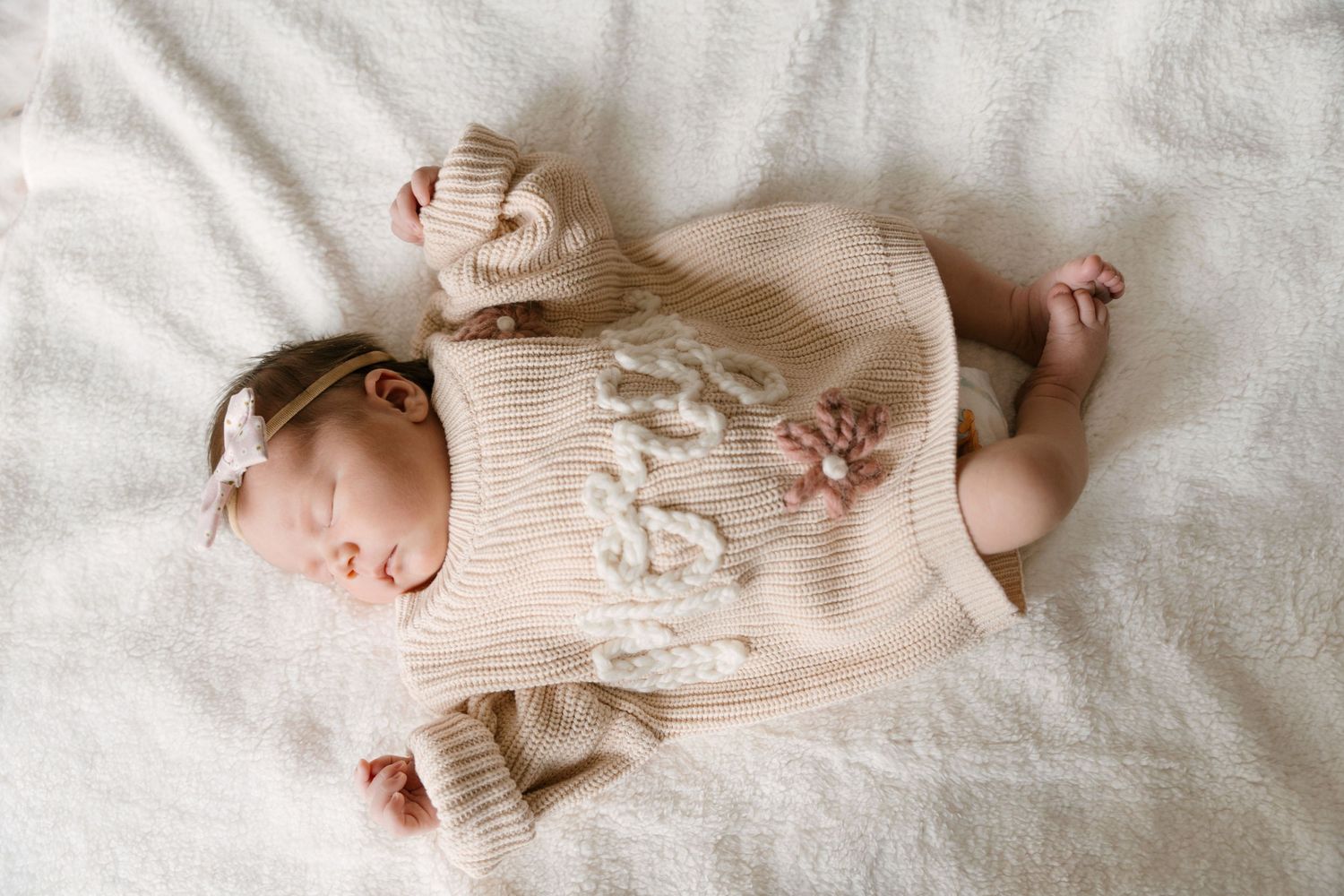 Sleeping newborn baby in cream knit sweater romper with bow headband on white blanket.