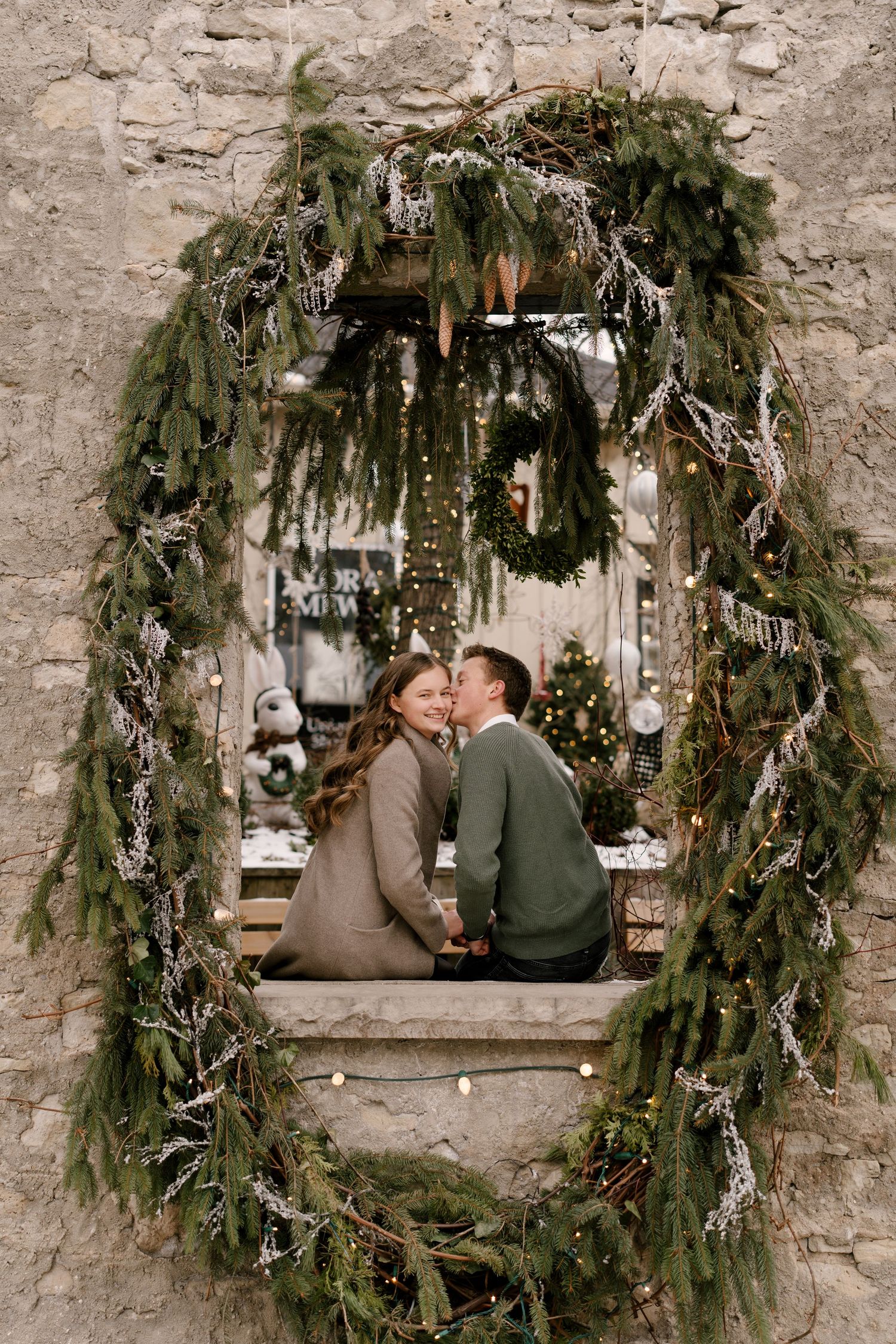 A romantic couple embraces inside a festive evergreen wreath arch with Christmas lights.