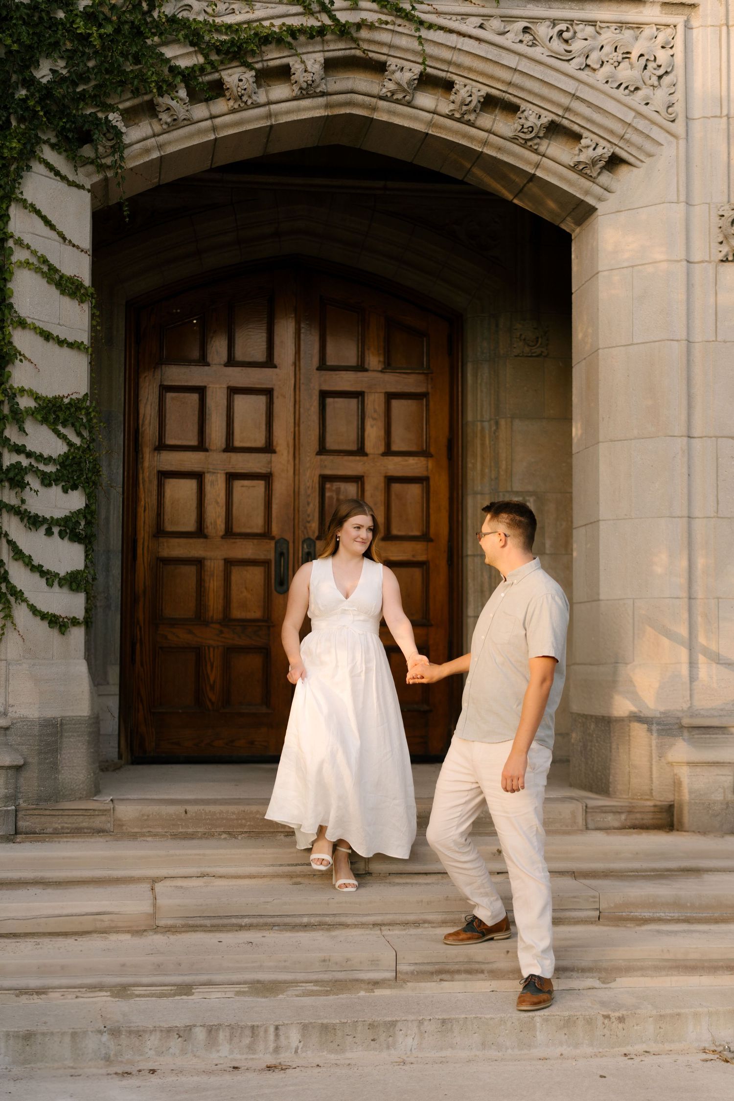Couple holding hands walking down stone steps of ornate ivy-covered building at sunset.
