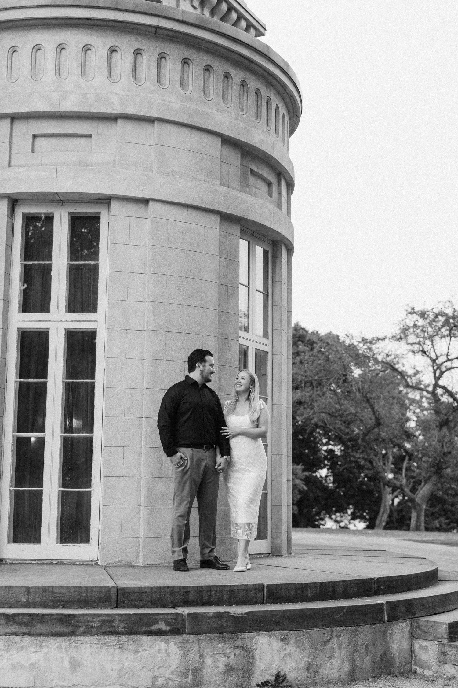 Black and white photo of elegant couple standing together beside a grand circular stone building.