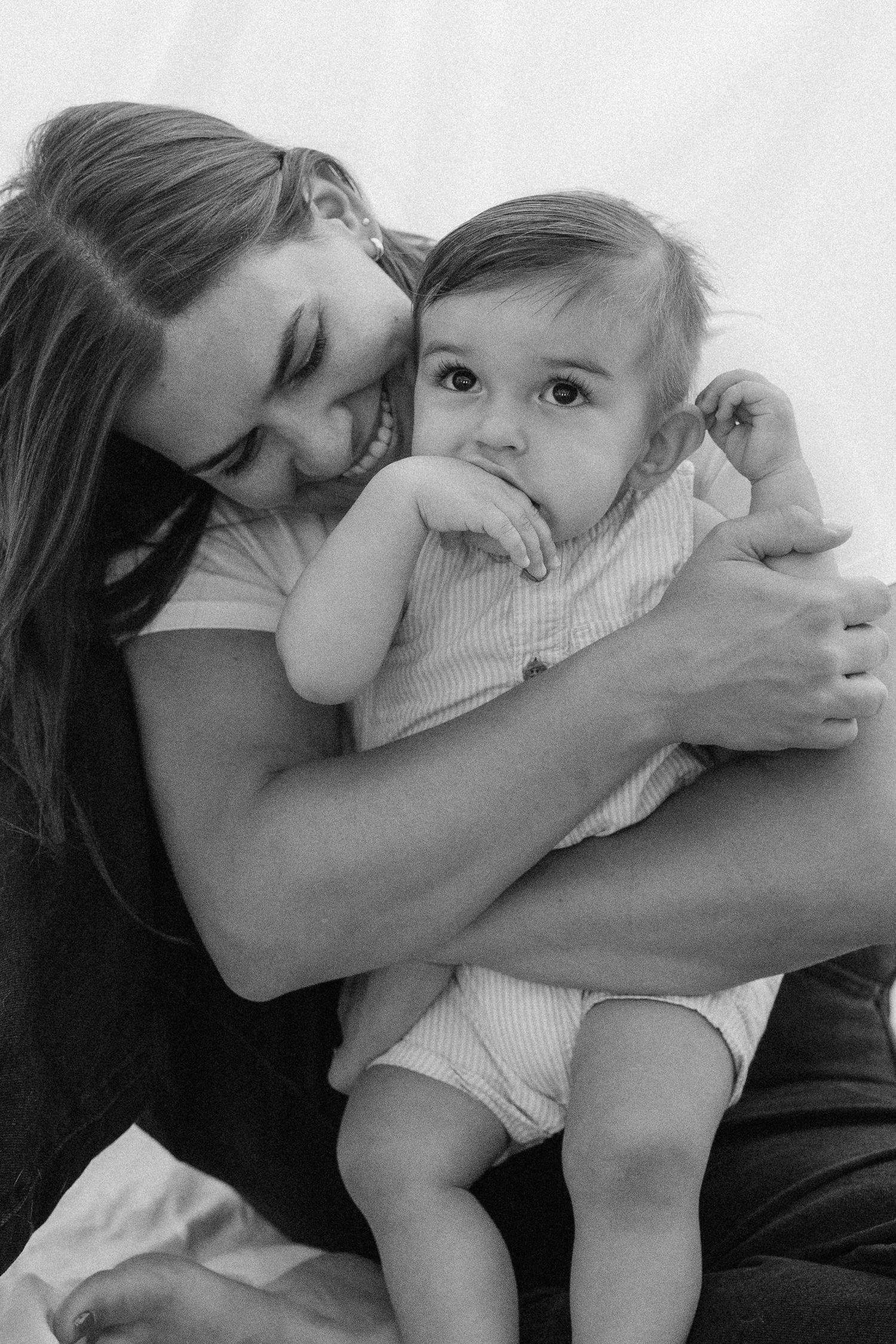 Black and white photo of a smiling mother tenderly holding her curious baby, sharing a sweet bonding moment.