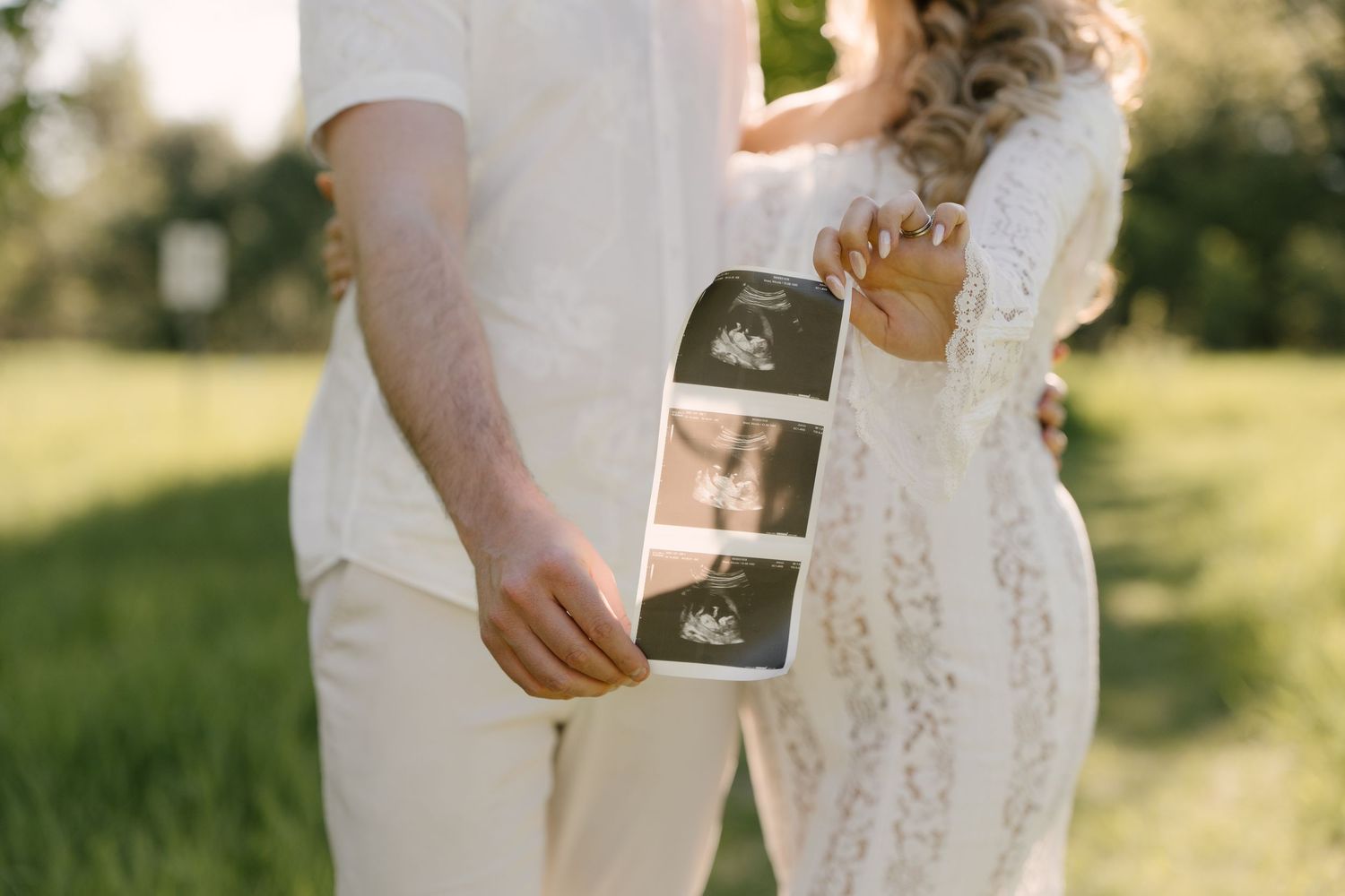 Expectant couple holding ultrasound photos outdoors in a sunny green field during maternity photoshoot.
