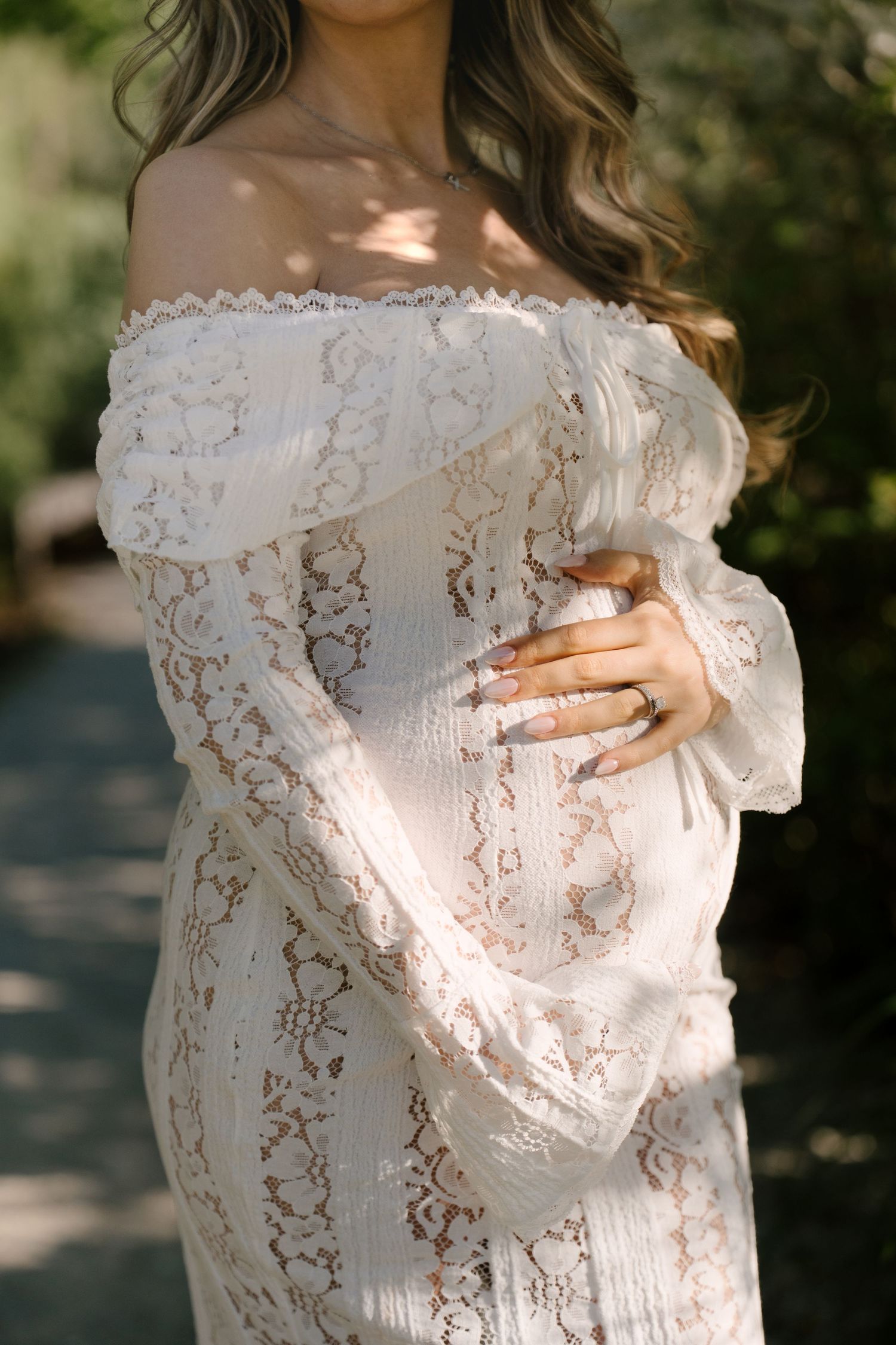 Close-up of pregnant woman in white off-shoulder lace dress gently cradling her baby bump.