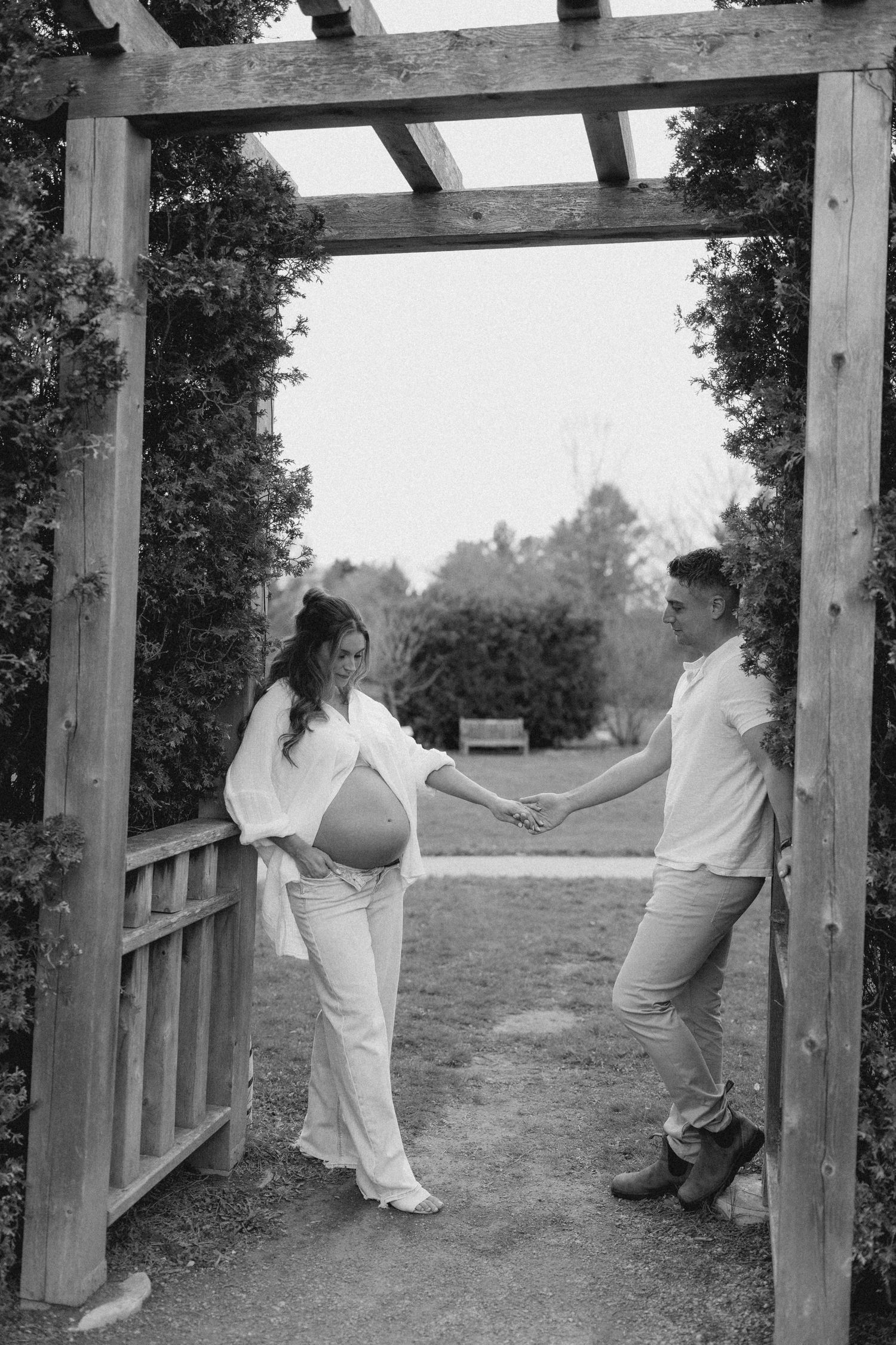 Black and white maternity photo of a pregnant woman and partner holding hands under a wooden pergola.