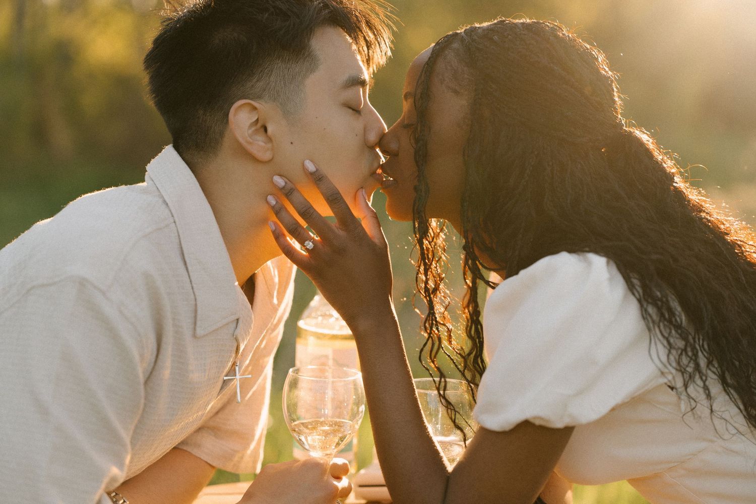 Interracial couple kissing outdoors holding wine glasses in warm sunset glow.