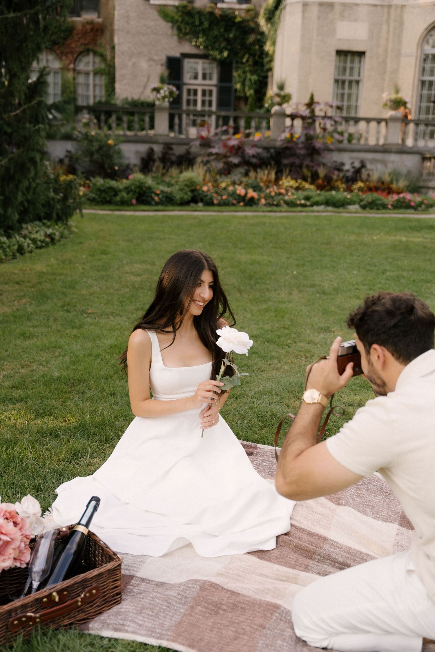 Man photographing smiling woman in white dress holding a white peony during garden picnic session.
