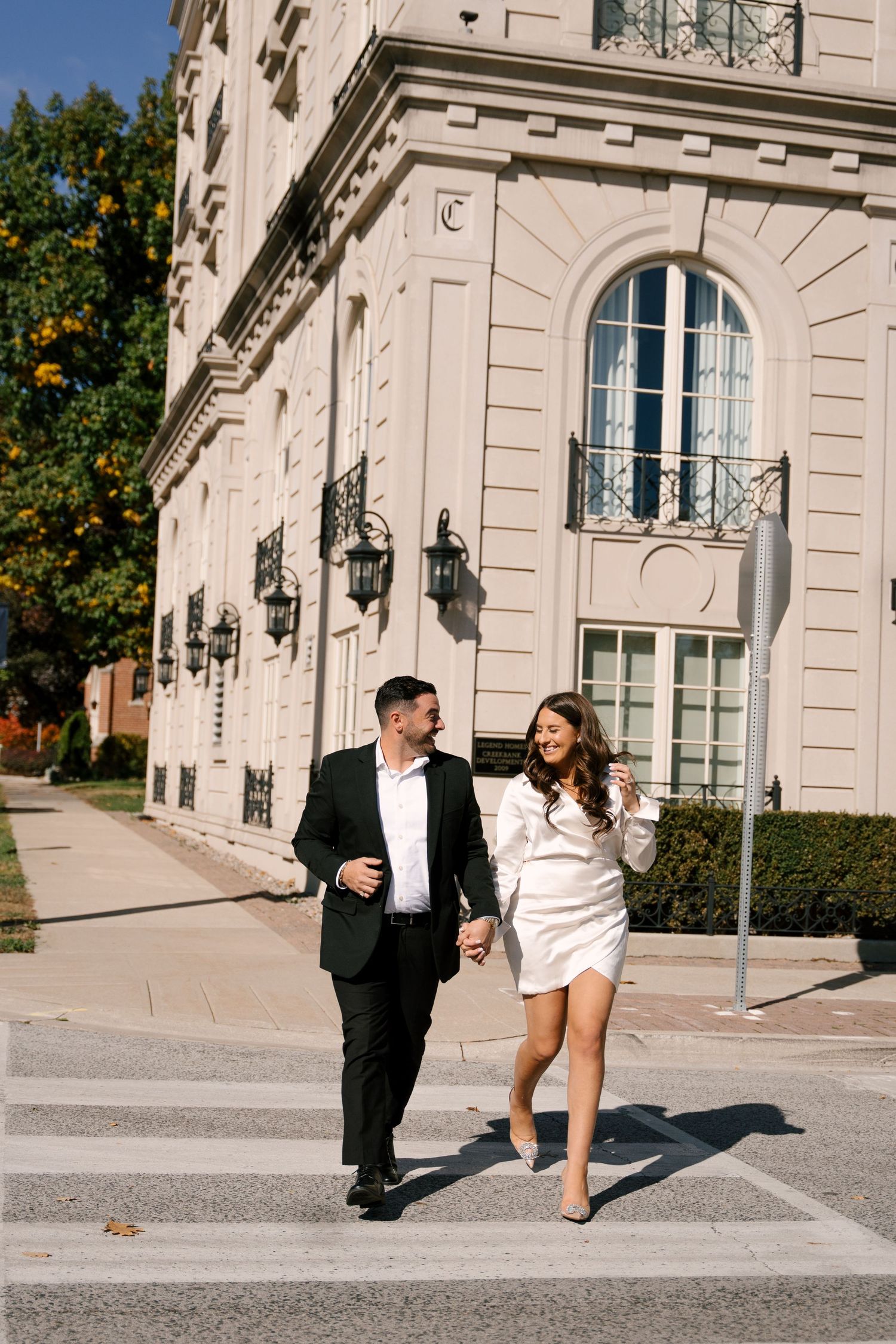 Happy engaged couple holding hands and walking across street near elegant white building outdoors.
