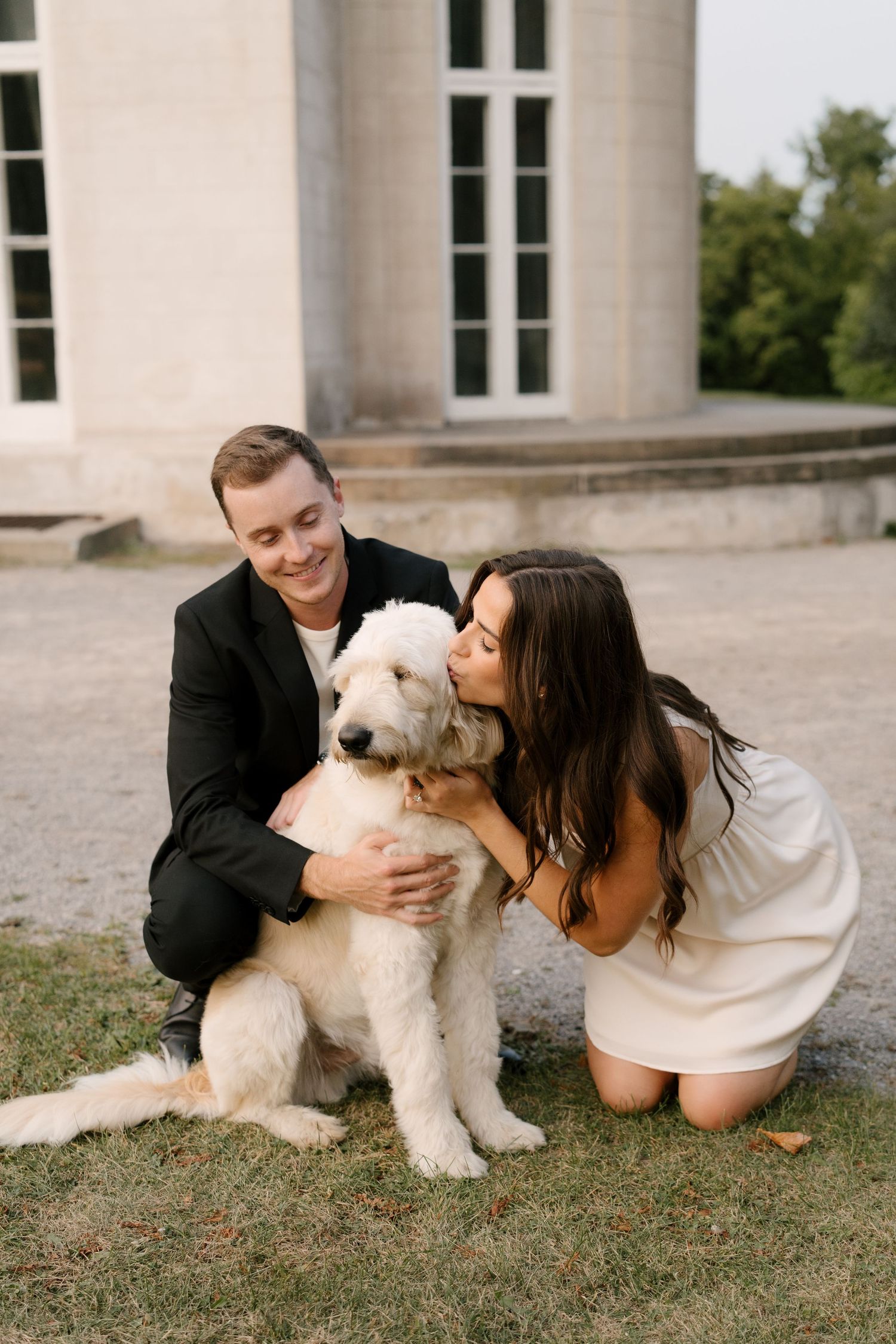 Couple crouching and kissing their fluffy white golden doodle dog outdoors near stone building.