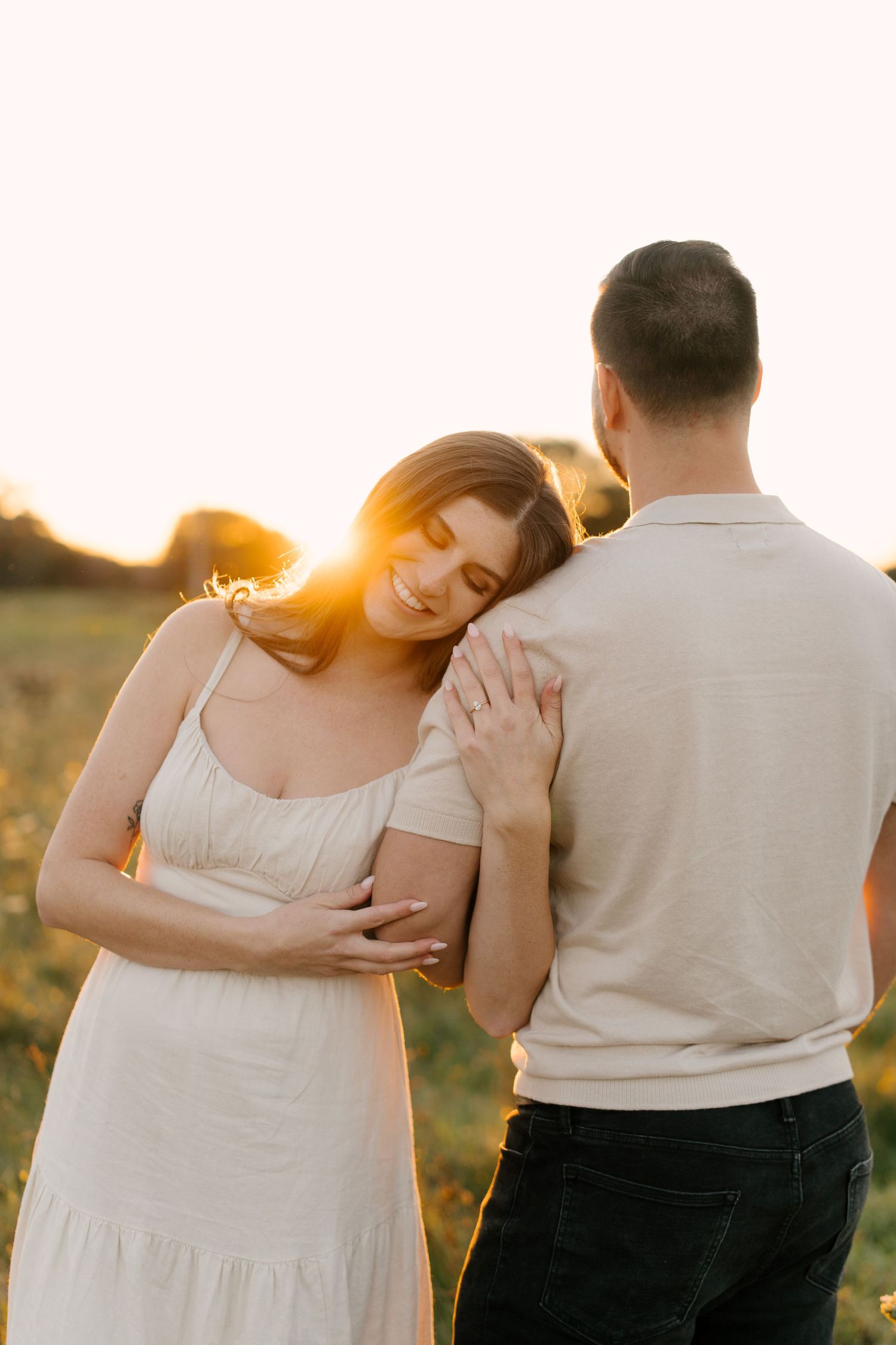 Woman leaning her head on her partners shoulder smiling softly in a warm sunset field.
