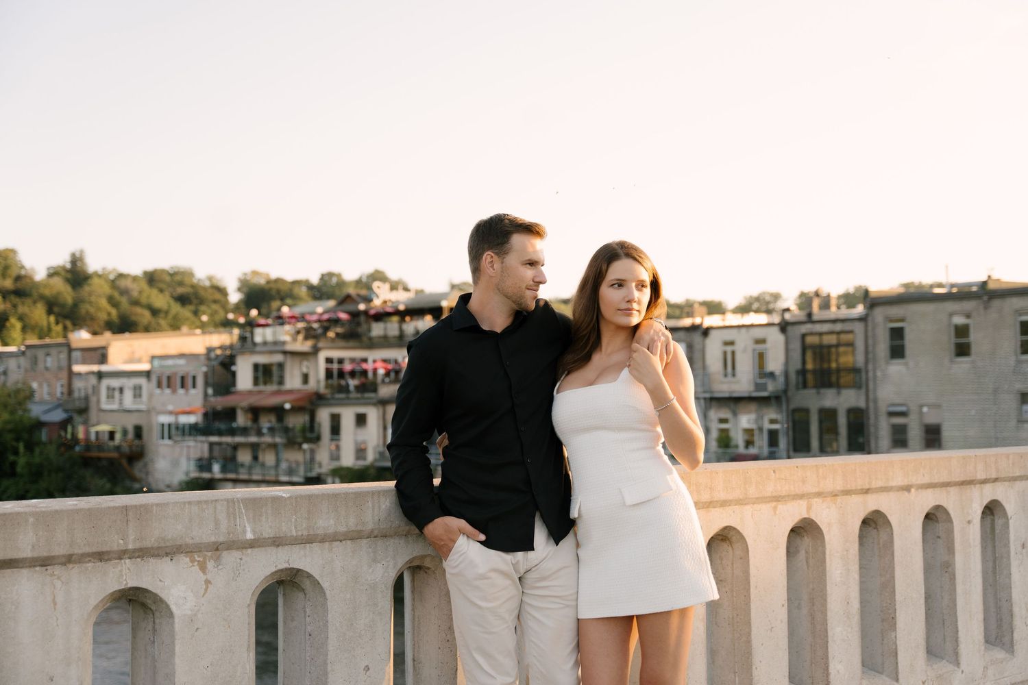Stylish couple standing on stone bridge overlooking charming town at sunset.