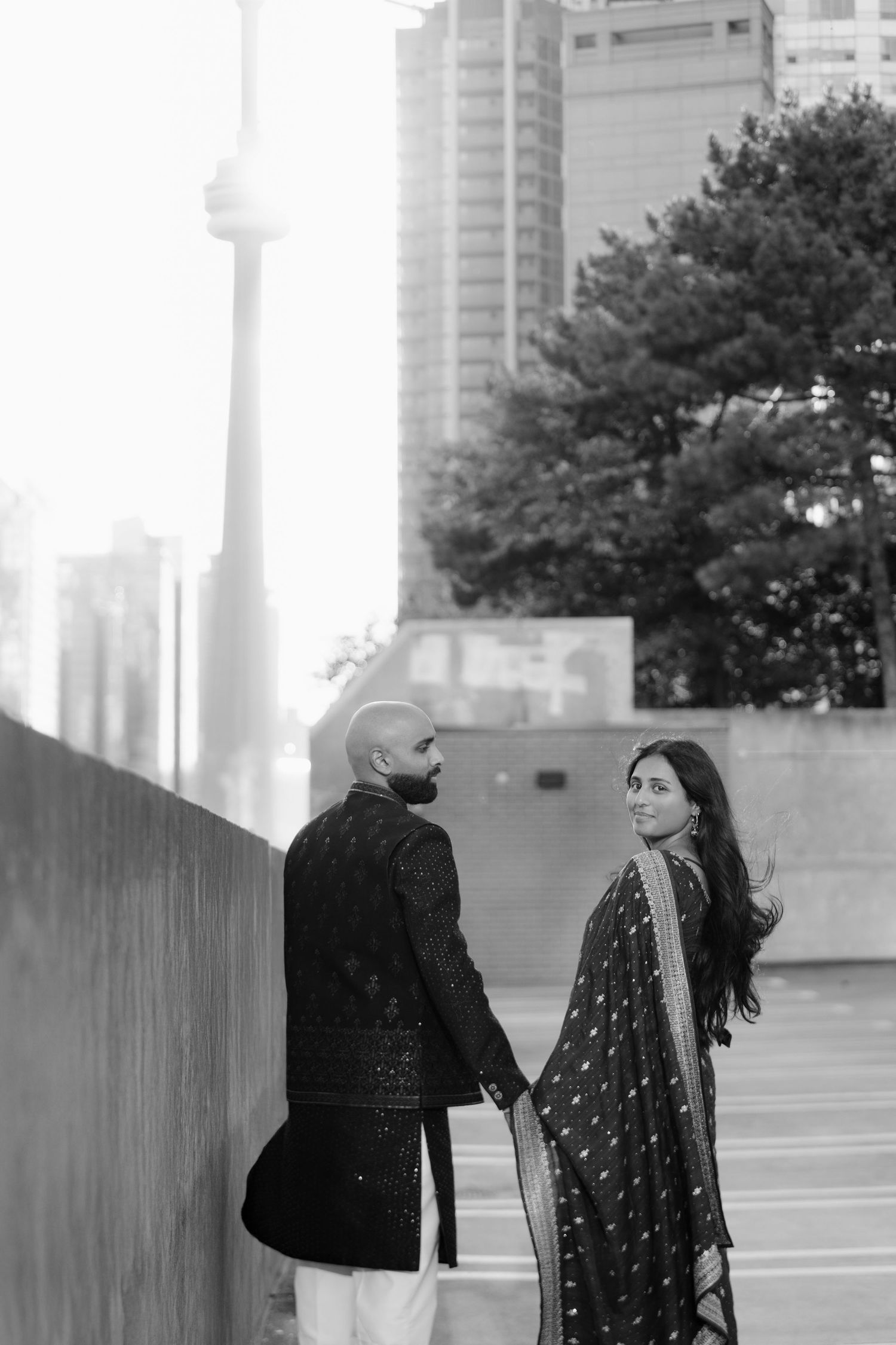 Couple in traditional South Asian attire holding hands with CN Tower in background, black and white in Toronto.
