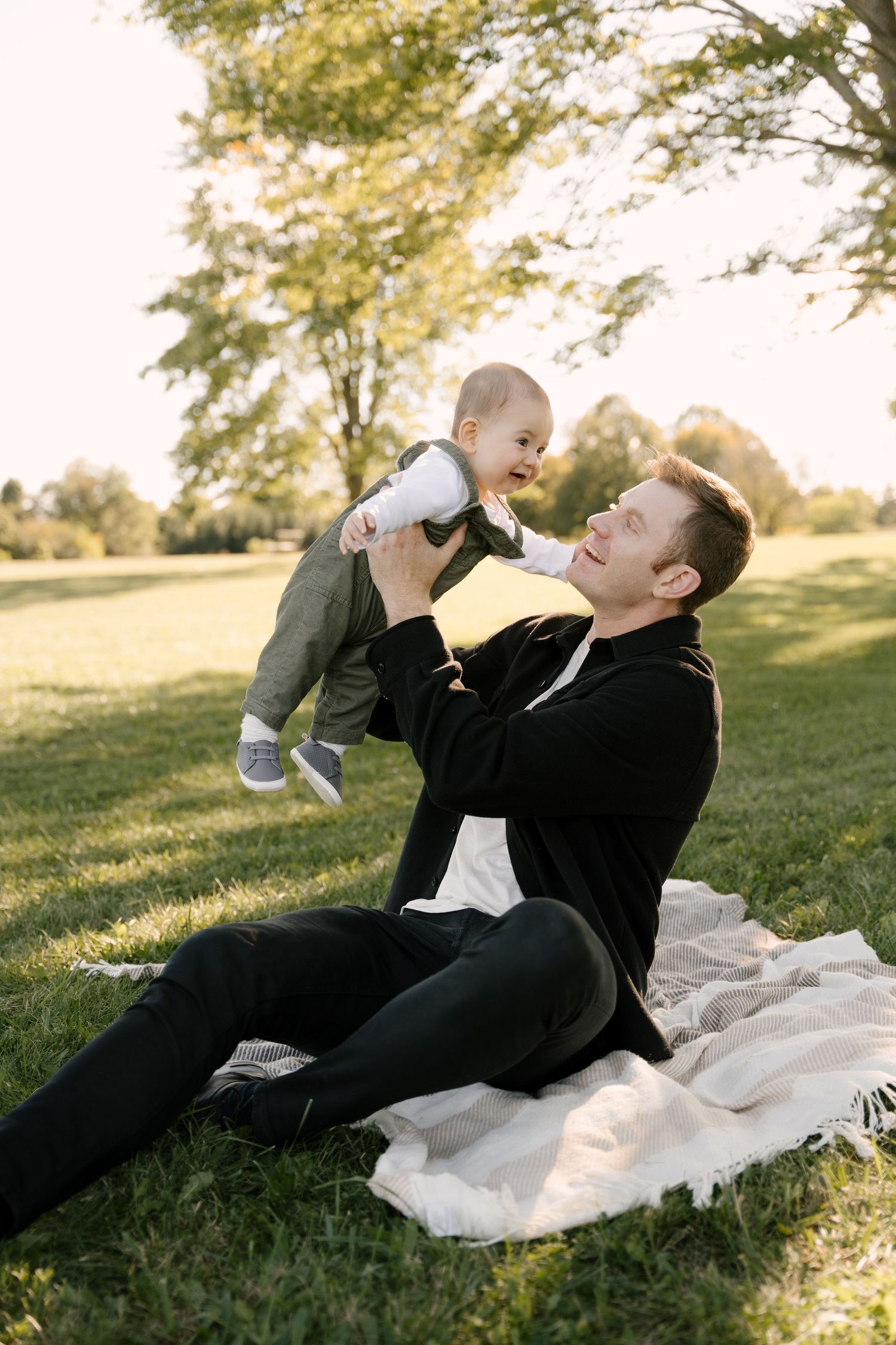 Father lifting laughing baby in park, sitting on blanket in golden sunlight.