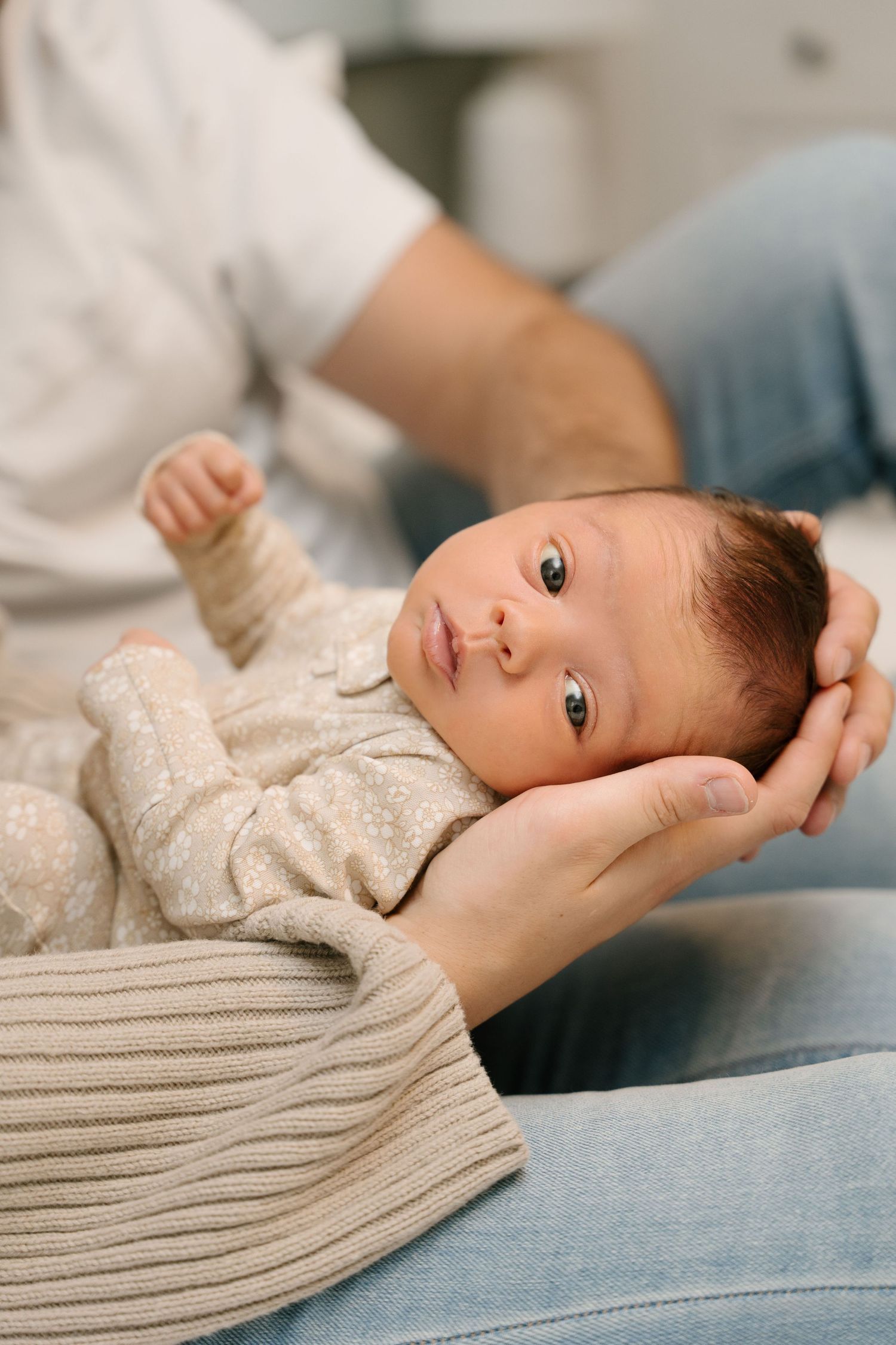 A newborn baby with dark hair lies alert in a parent's hands, wearing a beige floral onesie.