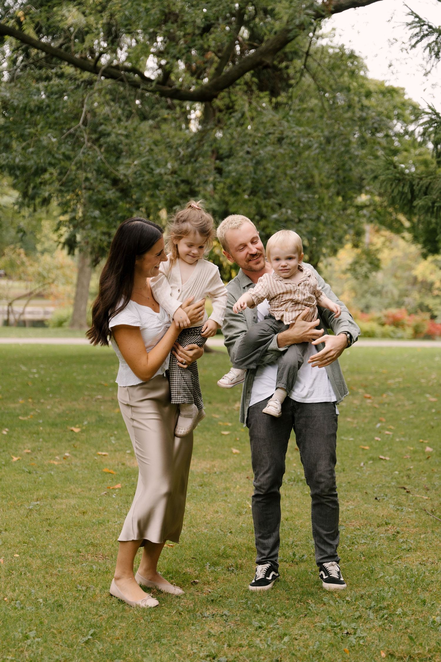 Family of four in park, parents holding two young children on green grass.